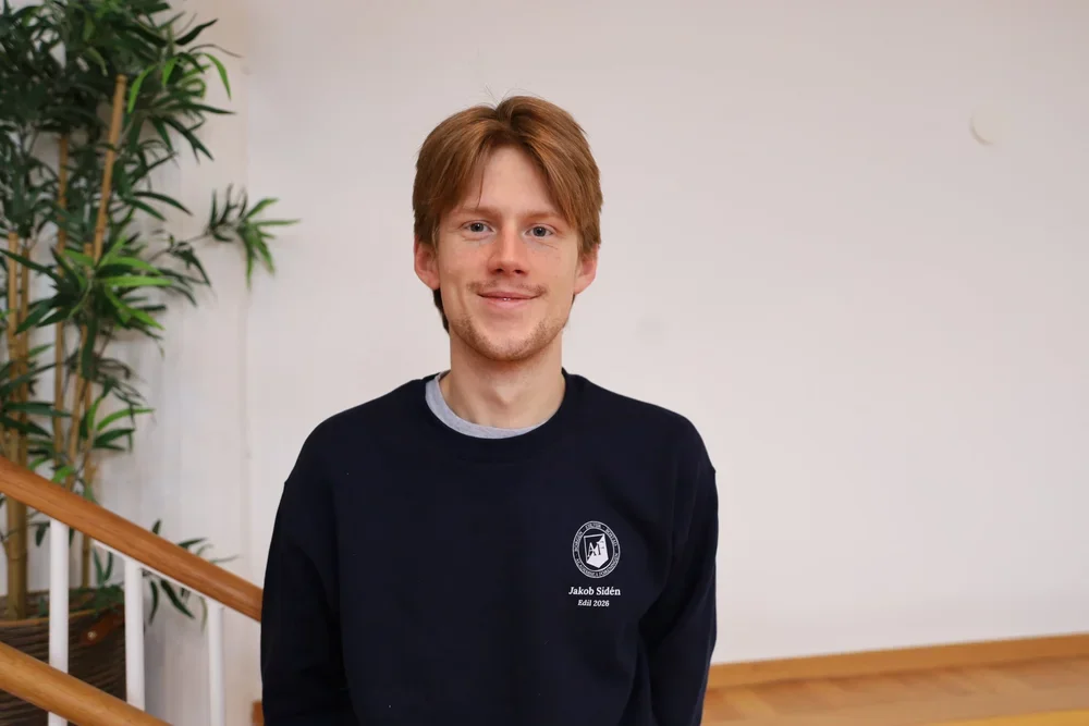 En ung man med rött hår och ljus hy, iklädd svart sweatshirt, står inomhus nära en trapp med ett grönt växt till vänster.