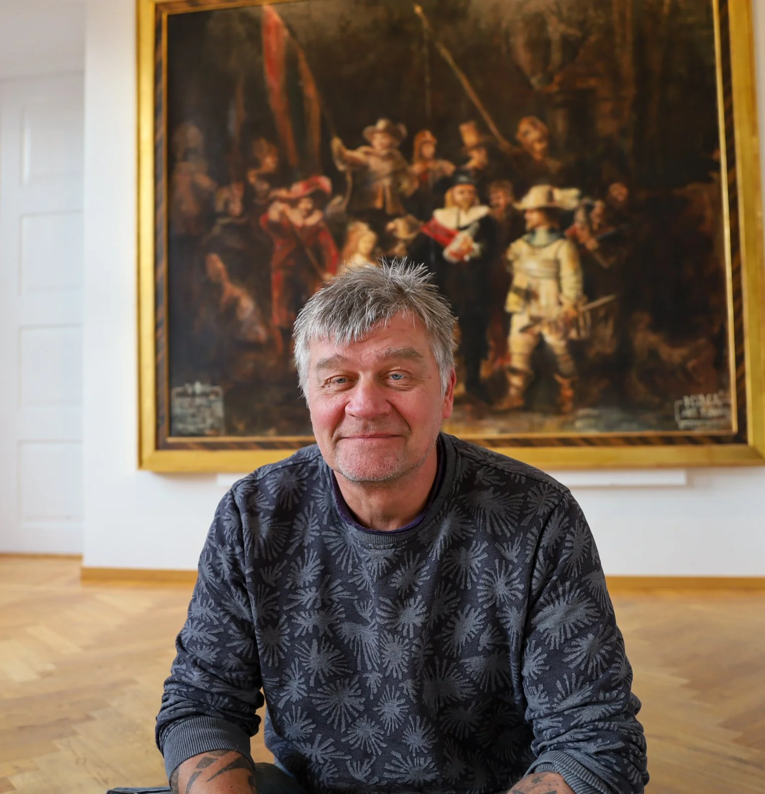 En man med grått hår och tatueringar på armen, sittande på golvet i ett konstgalleri, framför ett stort klassiskt konstverk i en guldförsedd ram.