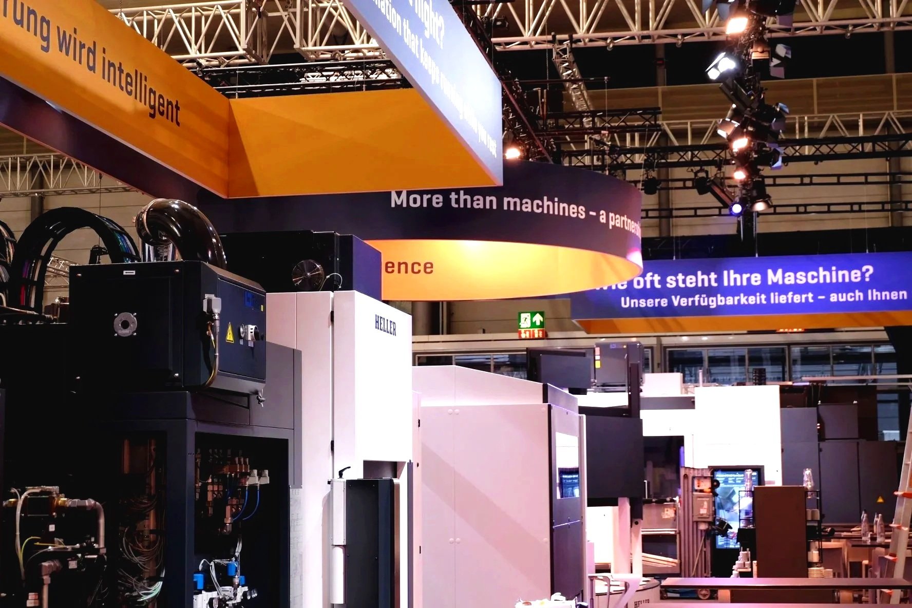 Exhibition booth with industrial machinery and large banners in orange and blue with text in German and English.