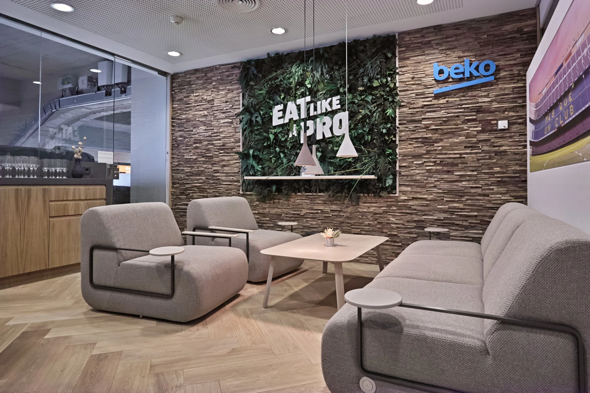 A modern lounge area with beige sofa and armchairs, wooden floor, a coffee table with a small flower arrangement, a stone accent wall with a green plant decor and the words "Eat Like a Pro" in white, and a blue Beko logo on the wall.