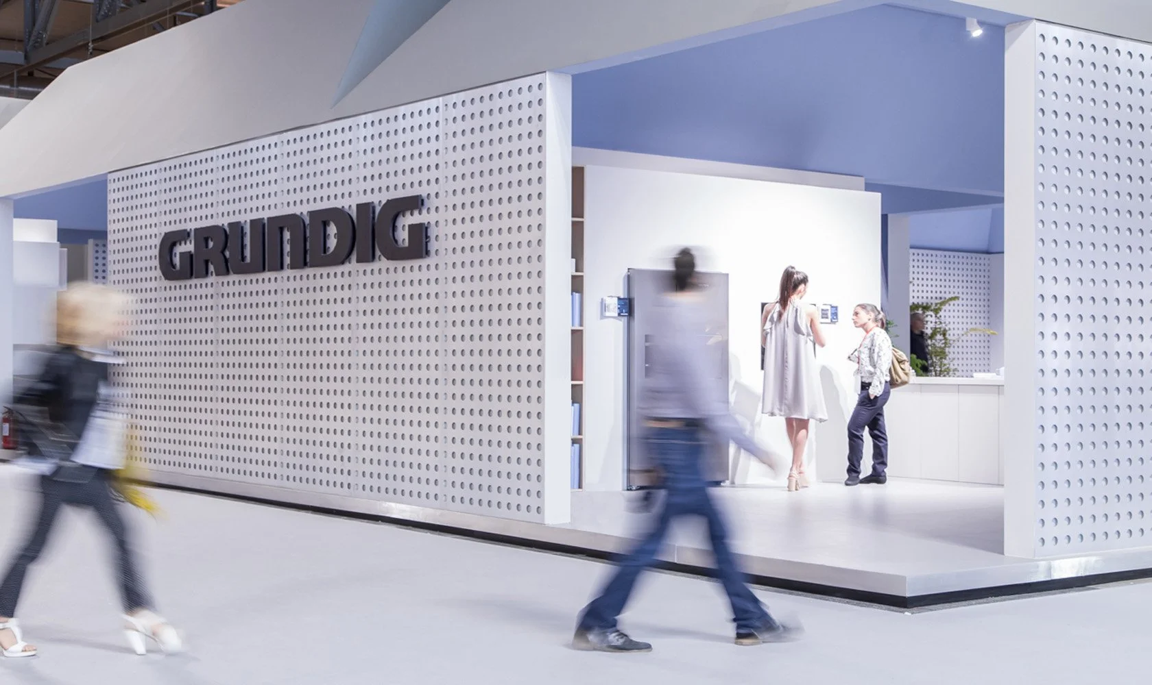 Exhibition booth with the brand name 'GRUNDIG' on a perforated white wall, featuring a reception counter and people interacting inside and outside the booth.