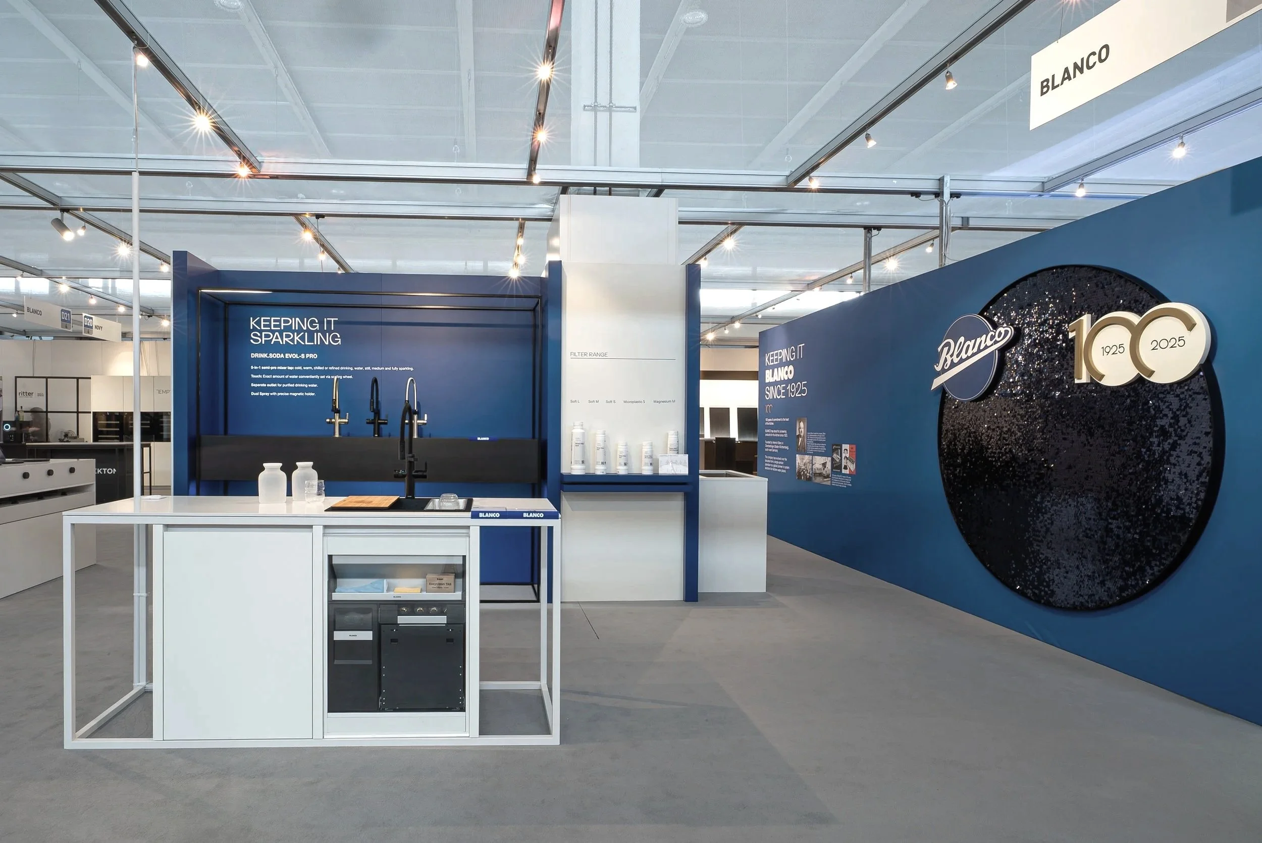Exhibition booth with blue and white color scheme, featuring kitchen fixtures and promotional signage for Blanco celebrating 100 years, 1925-2025.