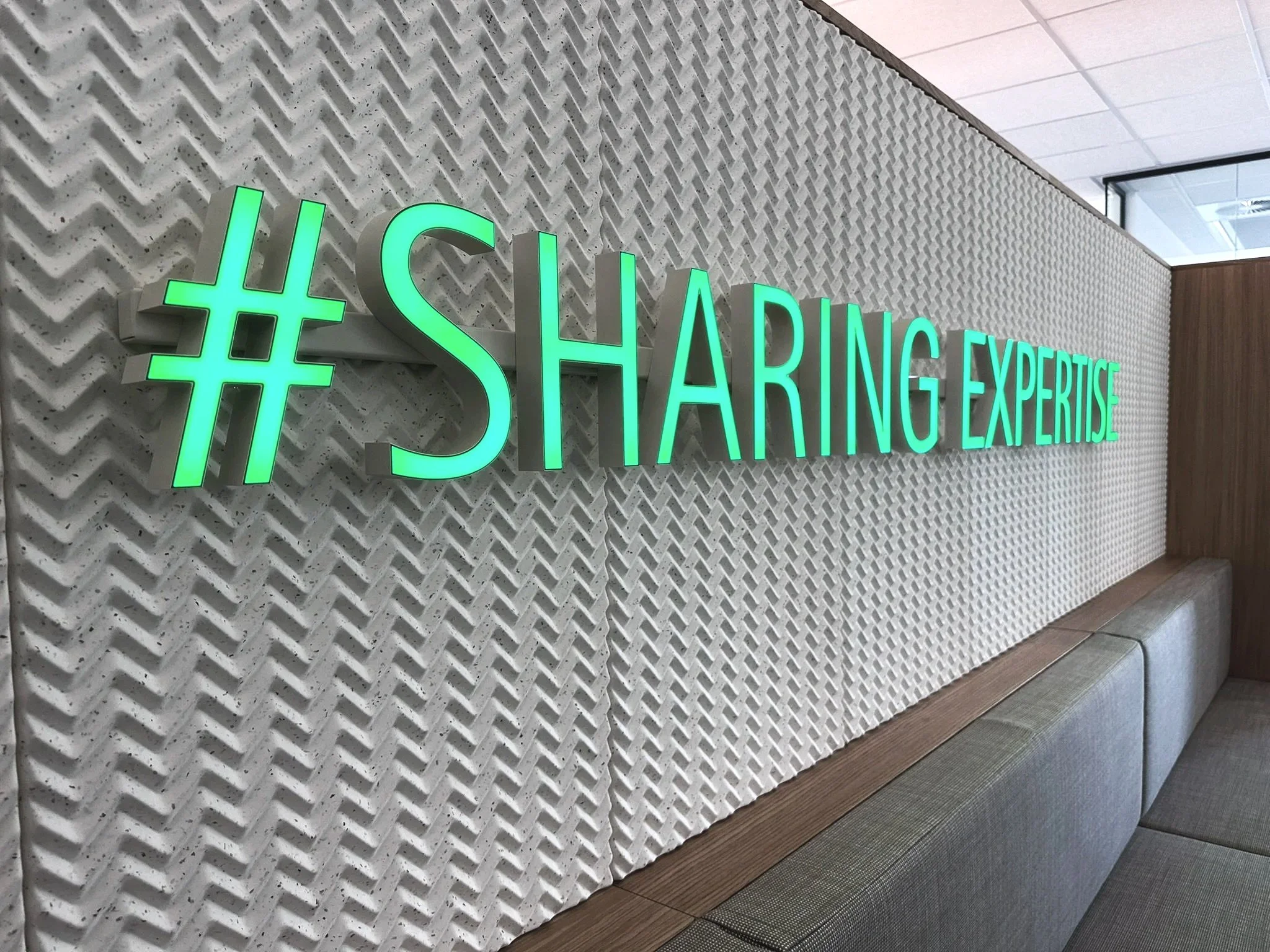 Neon sign reading '#SHARING EXPERTISE' in green, mounted on a textured white wall in an office.