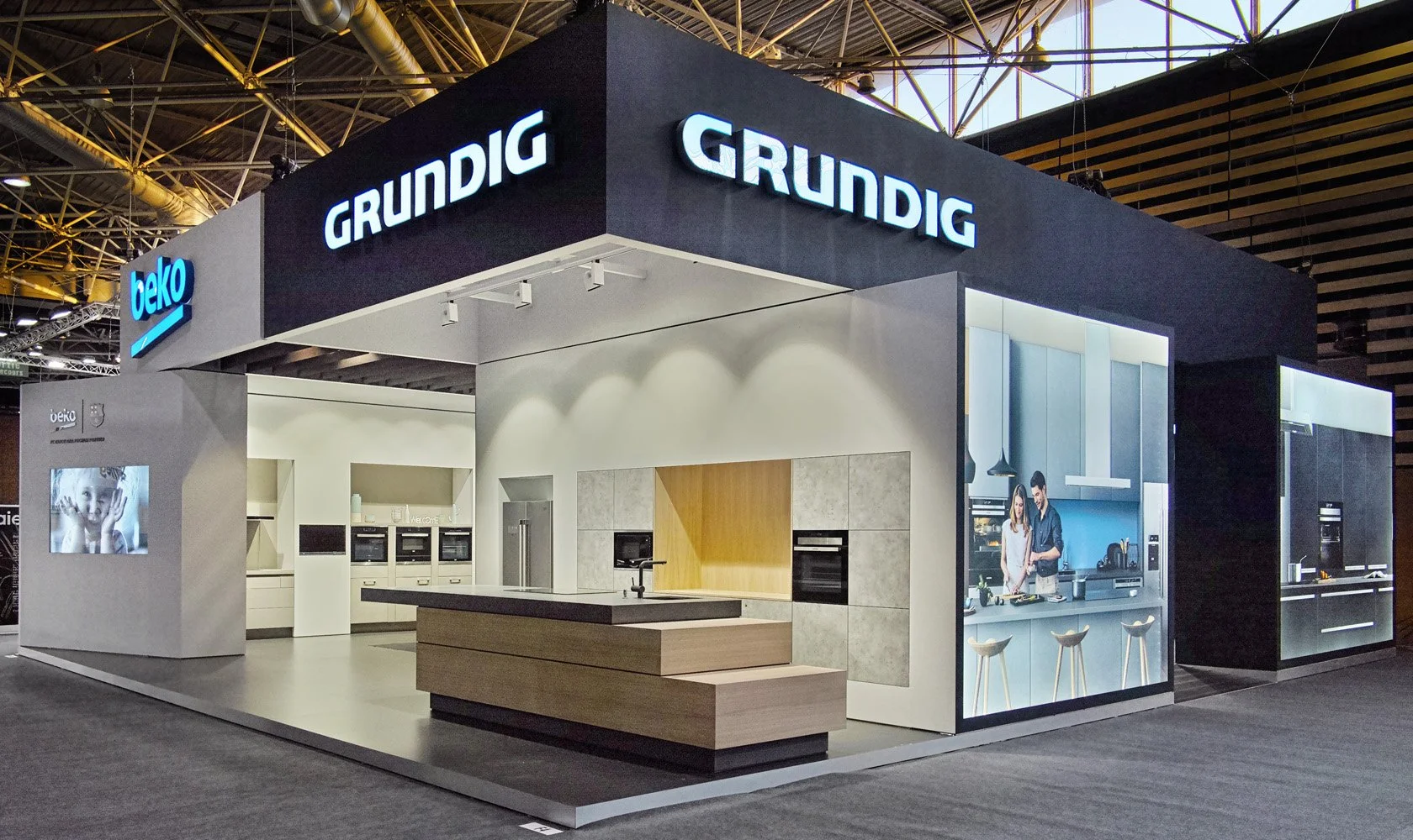 Trade show booth featuring brands Grundig, Beko, and Gorenje, showcasing kitchen appliances and cabinetry with models demonstrating devices in a modern kitchen setup.