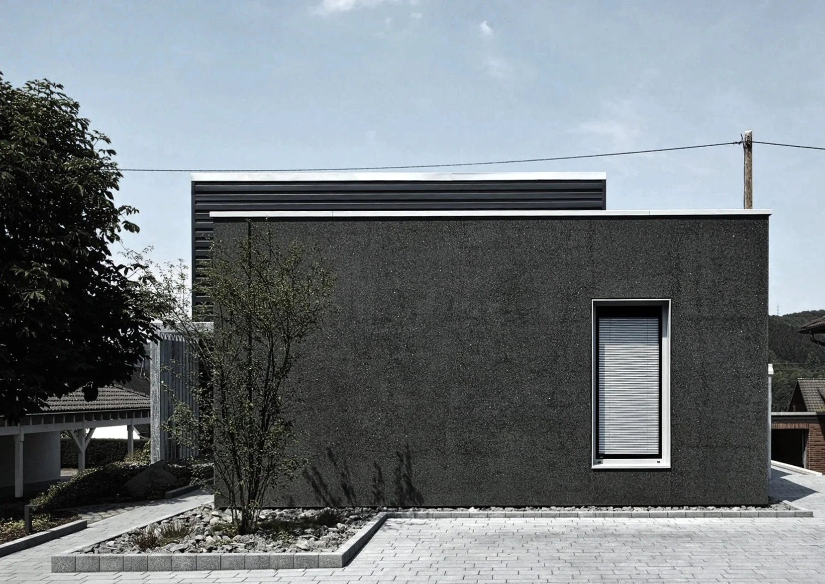 Modern black building with a single vertical window with blinds, small tree in front, paved ground, and a clear sky.