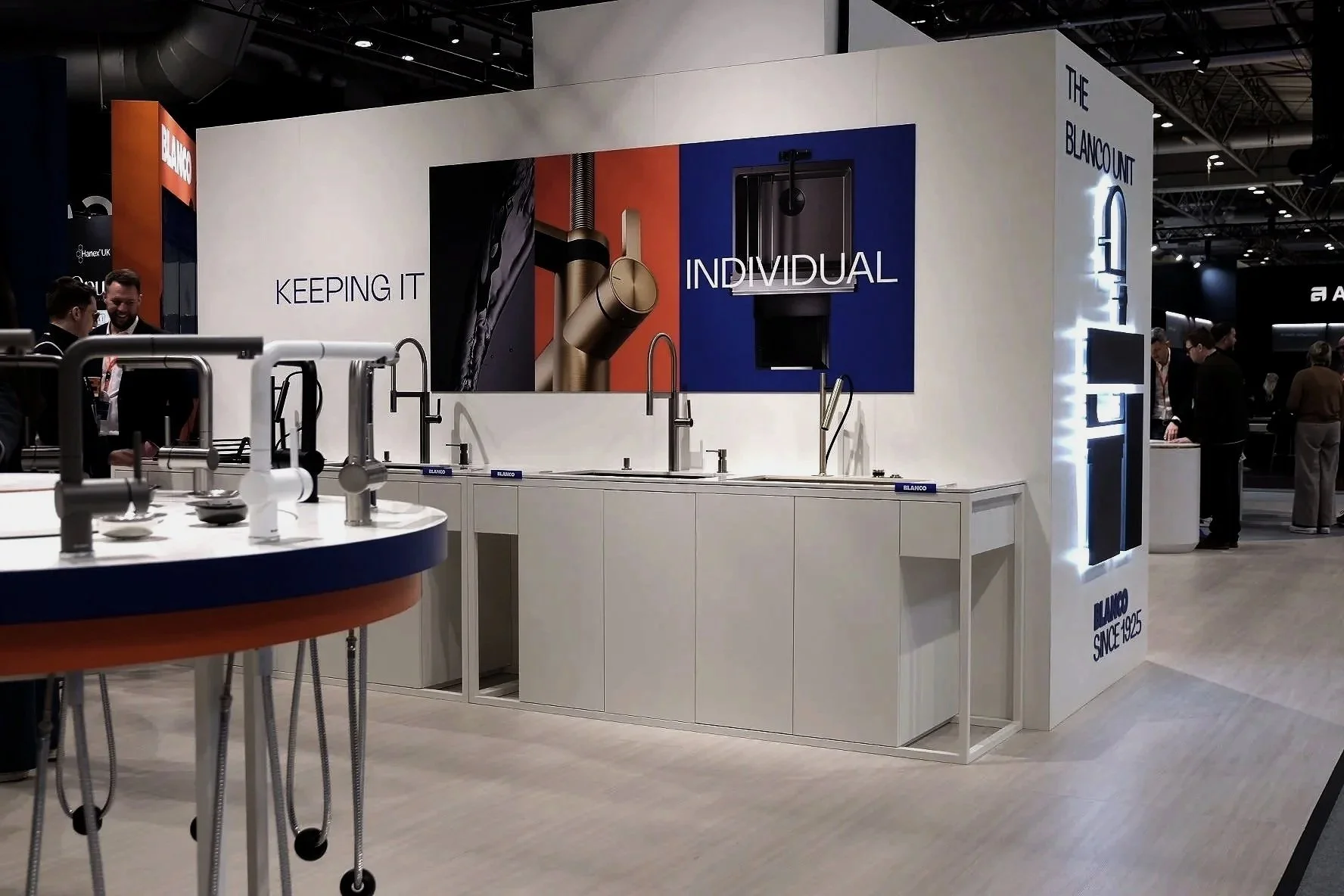 Exhibition booth showcasing kitchen faucets with branding 'Blanco' and slogan 'Keeping it Individual.' Several people are examining products, with a modern display of sinks and faucets in the foreground.