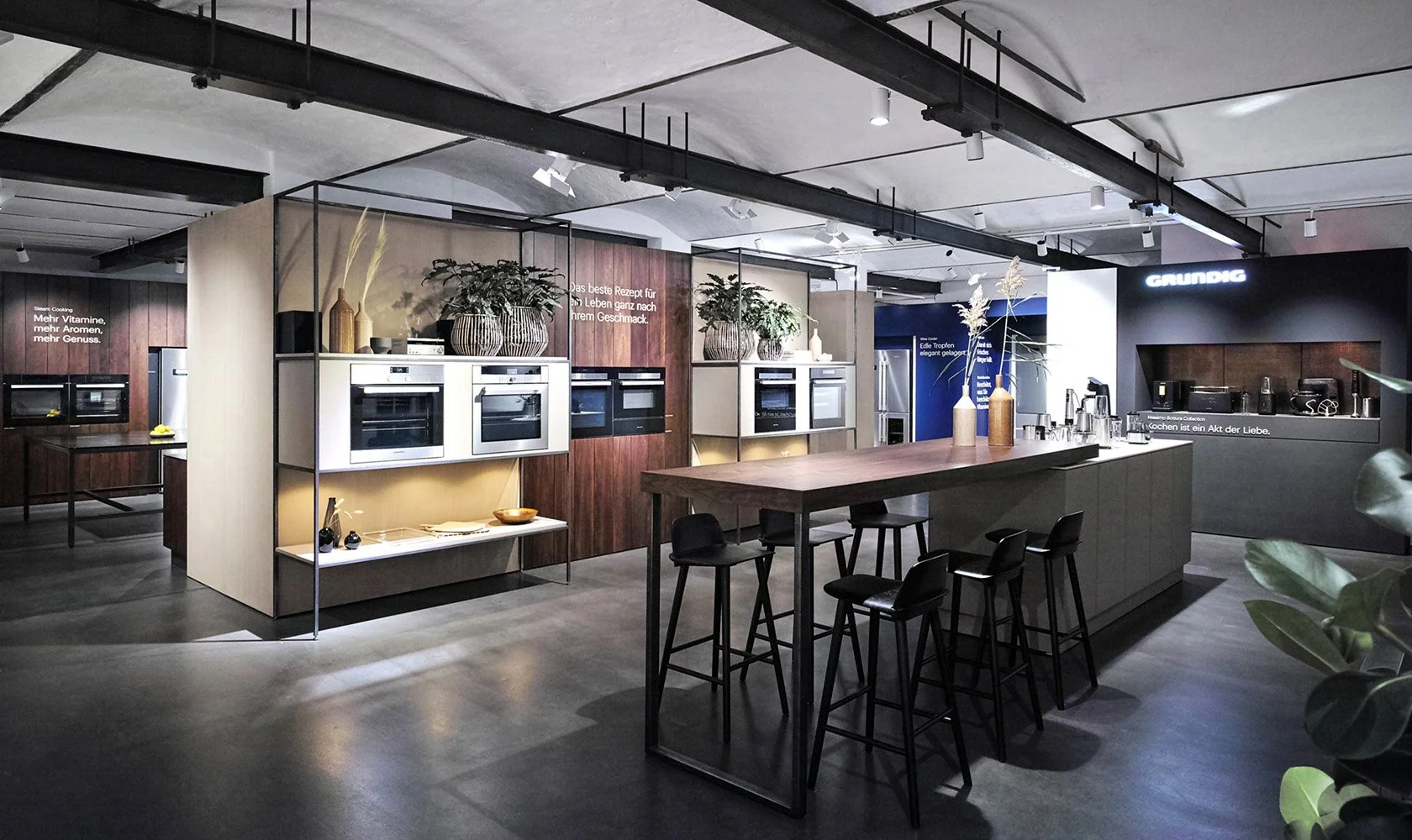 Modern kitchen showroom with wooden and white cabinets, built-in ovens, a large island with bar stools, decorative vases, and potted plants, under industrial style lighting.