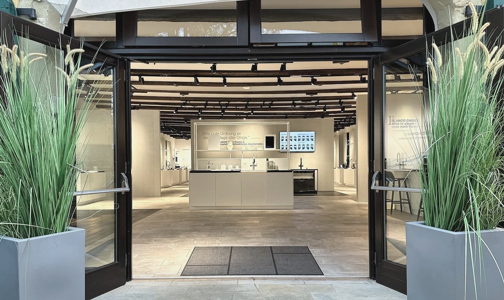 View of a modern kitchen showroom with an open entrance framed by large potted green plants on either side. Inside, there are white kitchen displays, faucets, and digital screens showcasing products.