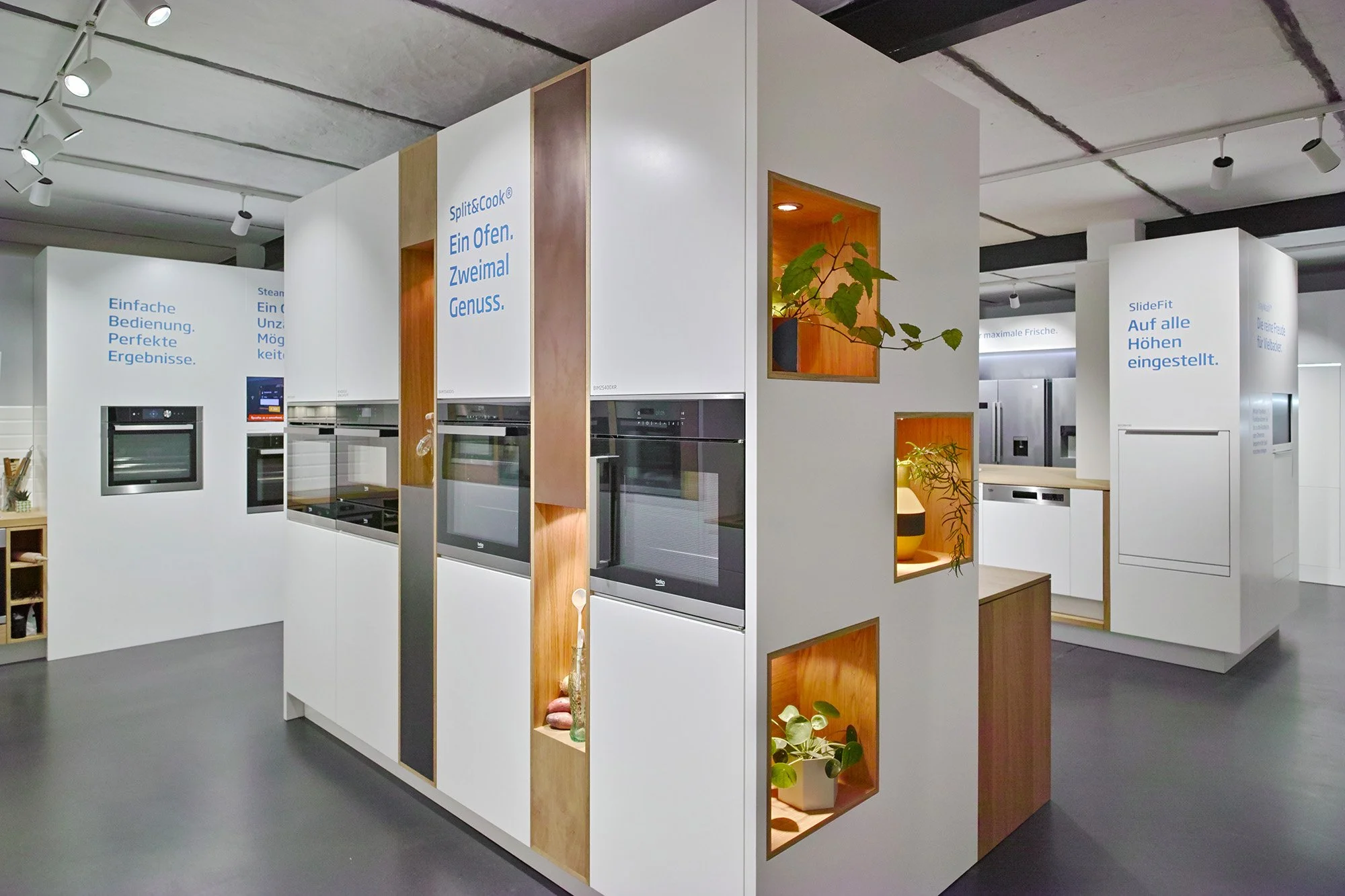 Showroom display of modern kitchen appliances, including built-in ovens and refrigerators, with informational signs in German and decorative plants on wooden shelves.
