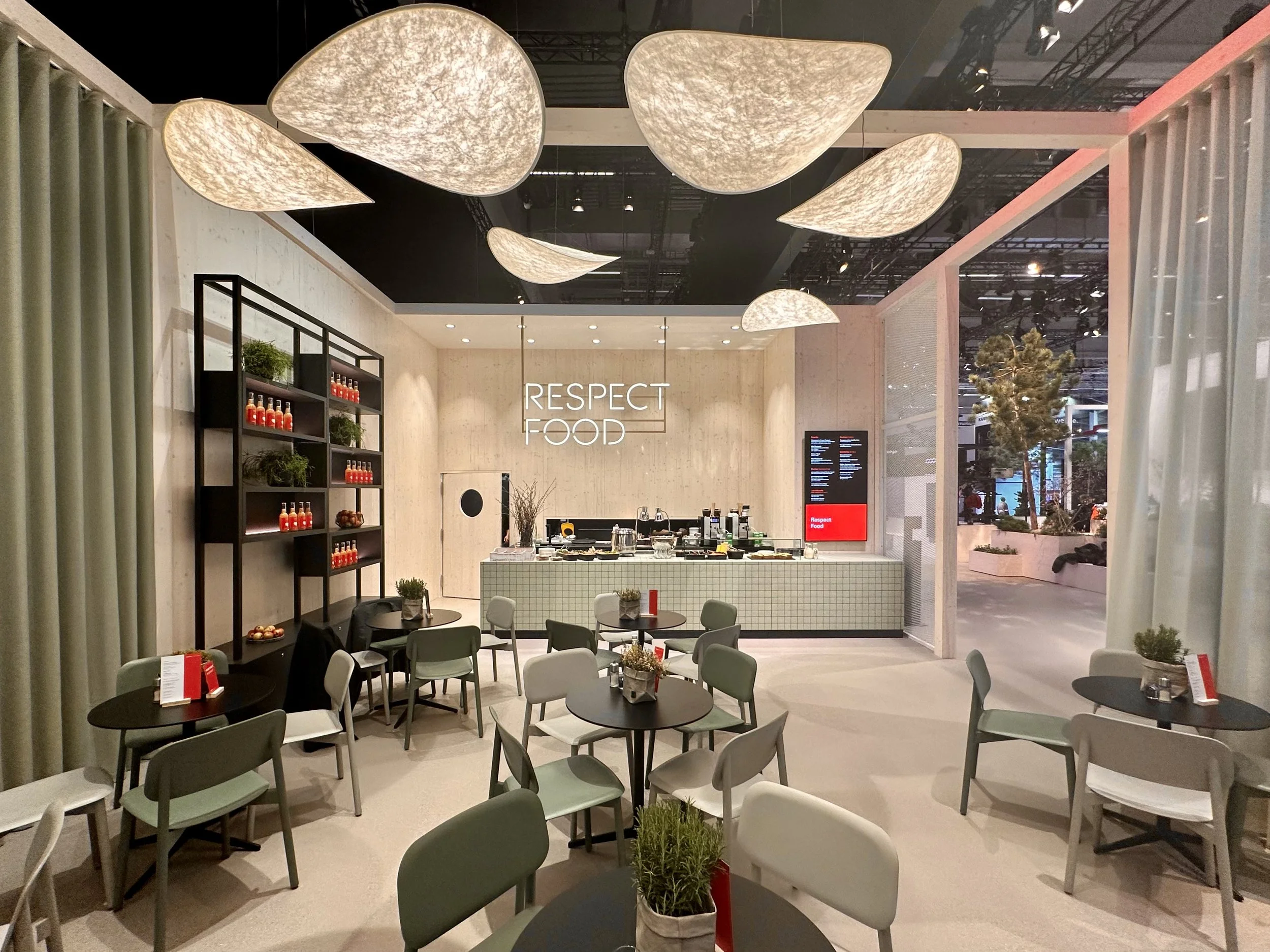 Modern dining area with pastel-colored chairs and round black tables, decorated with small plants. Behind is a counter with a hanging sign that says 'Respect Food,' and a display with supplies. Large, abstract ceiling lights hang above, and large windows reveal an outdoor seating area with trees.