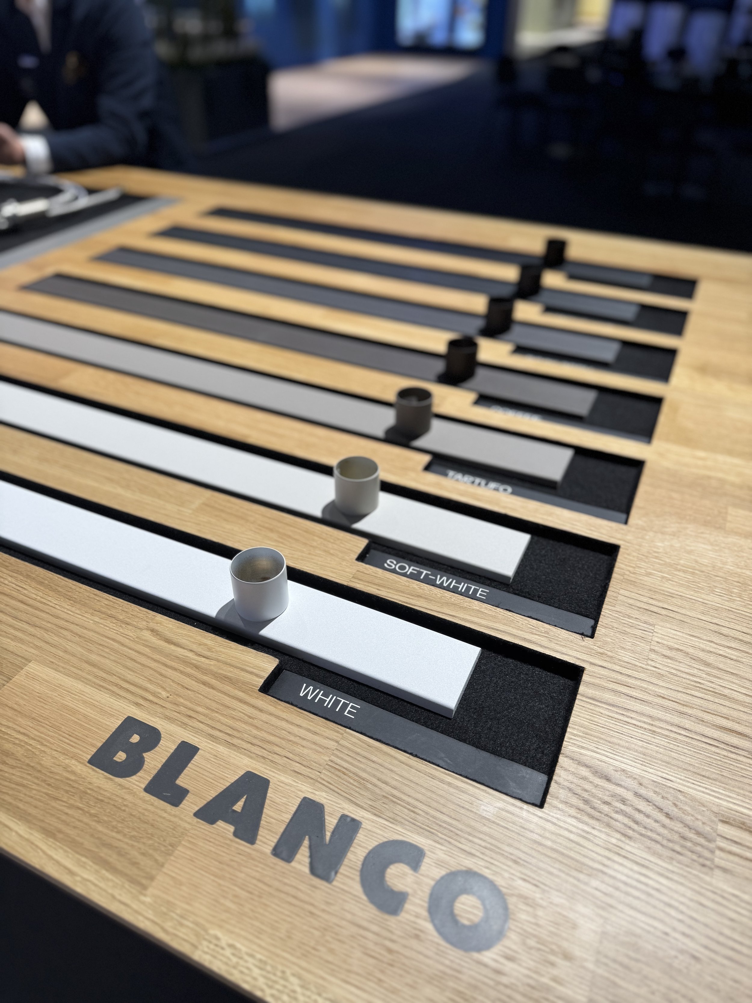 Close-up of a wooden display board with sliders showing different shades of black, gray, and white, labeled as 'WHITE', 'SOFT-WHITE', and 'BLACK'. The word 'BLANCO' is written on the bottom left corner of the board. The background shows a person in a suit sitting at a nearby table.