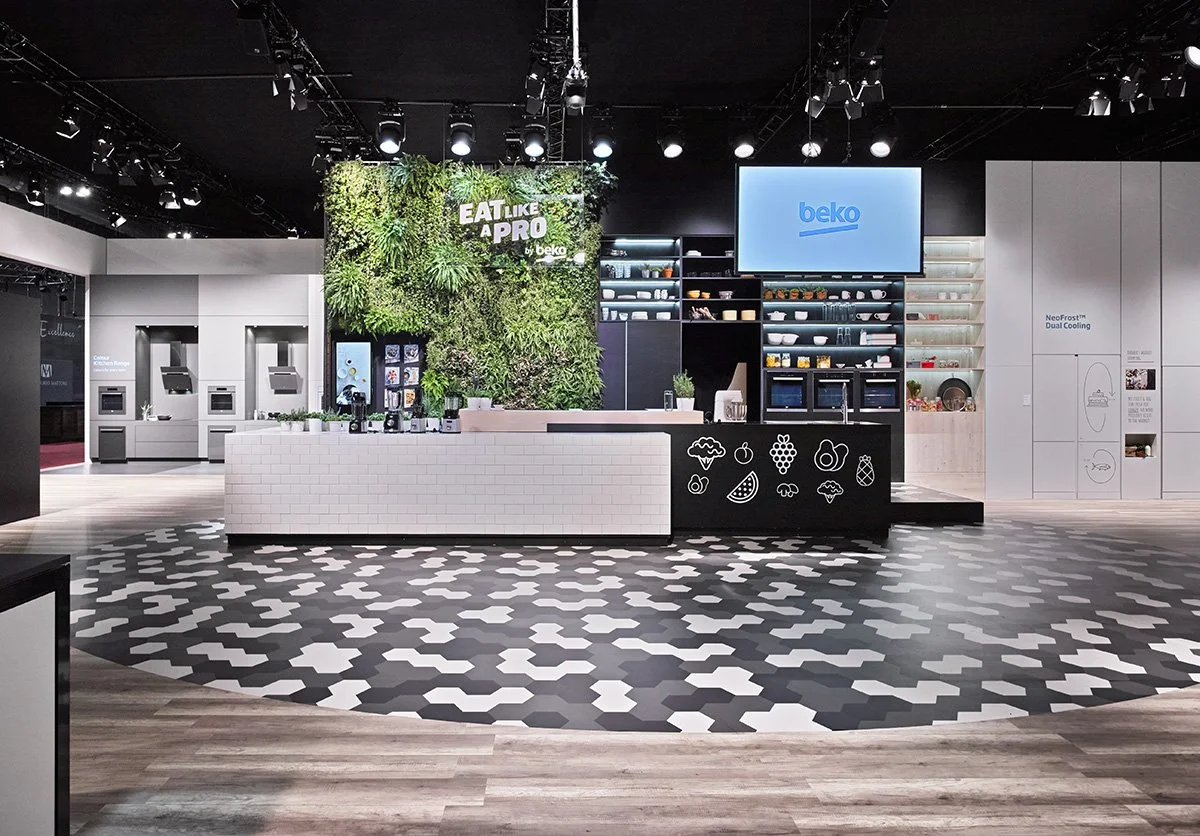 Display of a modern kitchen with a black and white tiled floor, white and black cabinetry, and a large green vertical garden wall with the phrase 'Eat Like a Pro by Beko' above.