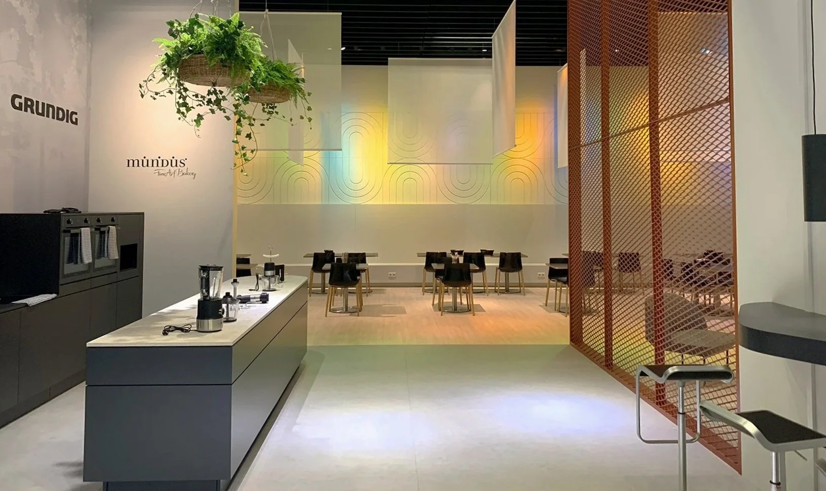 Modern cafe with a counter on the left, a hanging plant above, and a dining area with black chairs and small tables in the background, illuminated by multicolored lighting on the wall.