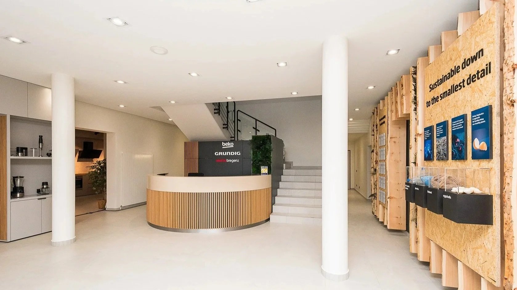 Modern office reception area with white walls, wooden accents, a curved reception desk, and informational display boards about sustainability.