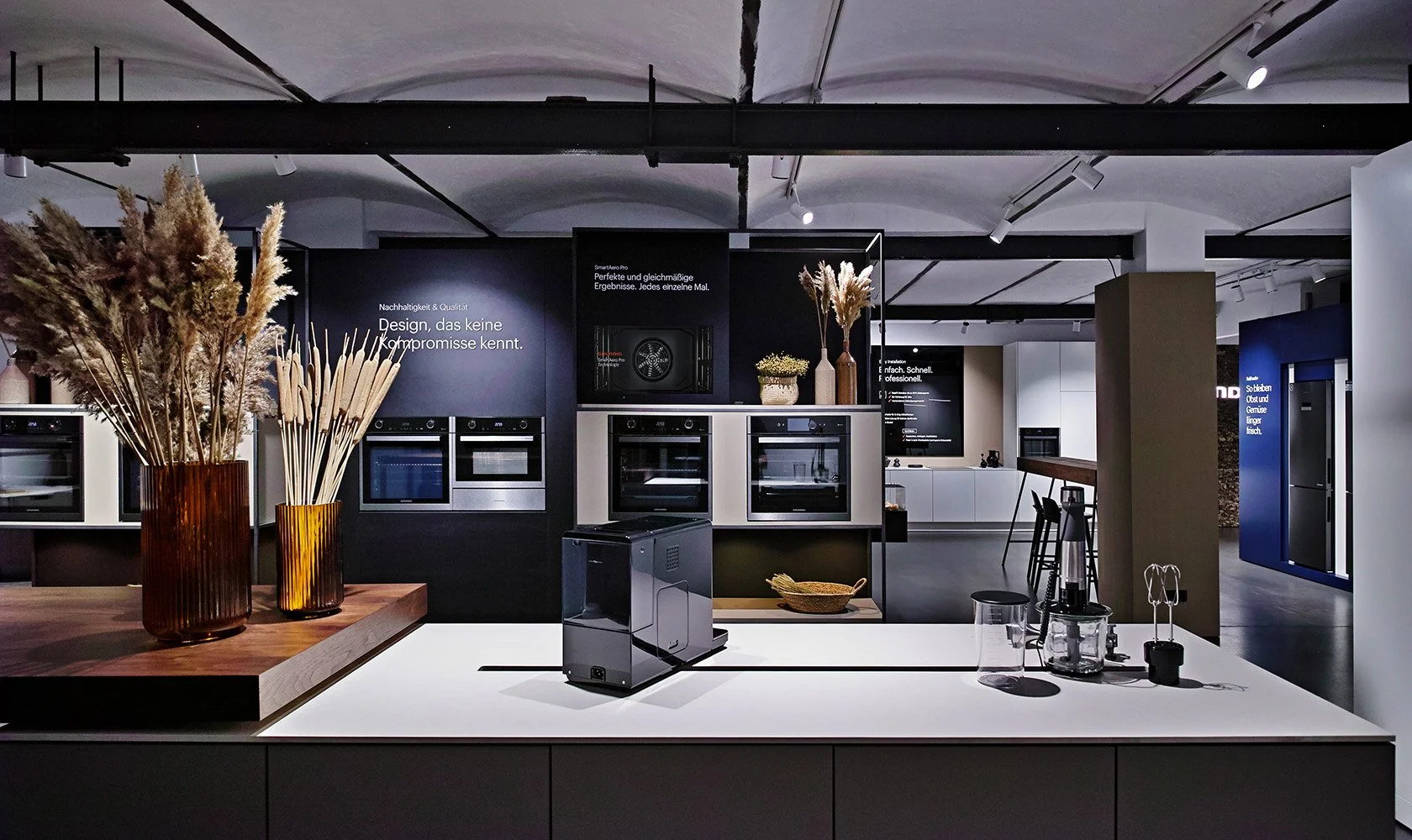 Display of modern kitchen appliances and decor in a showroom, including ovens, vases with dried flowers, and kitchen gadgets.