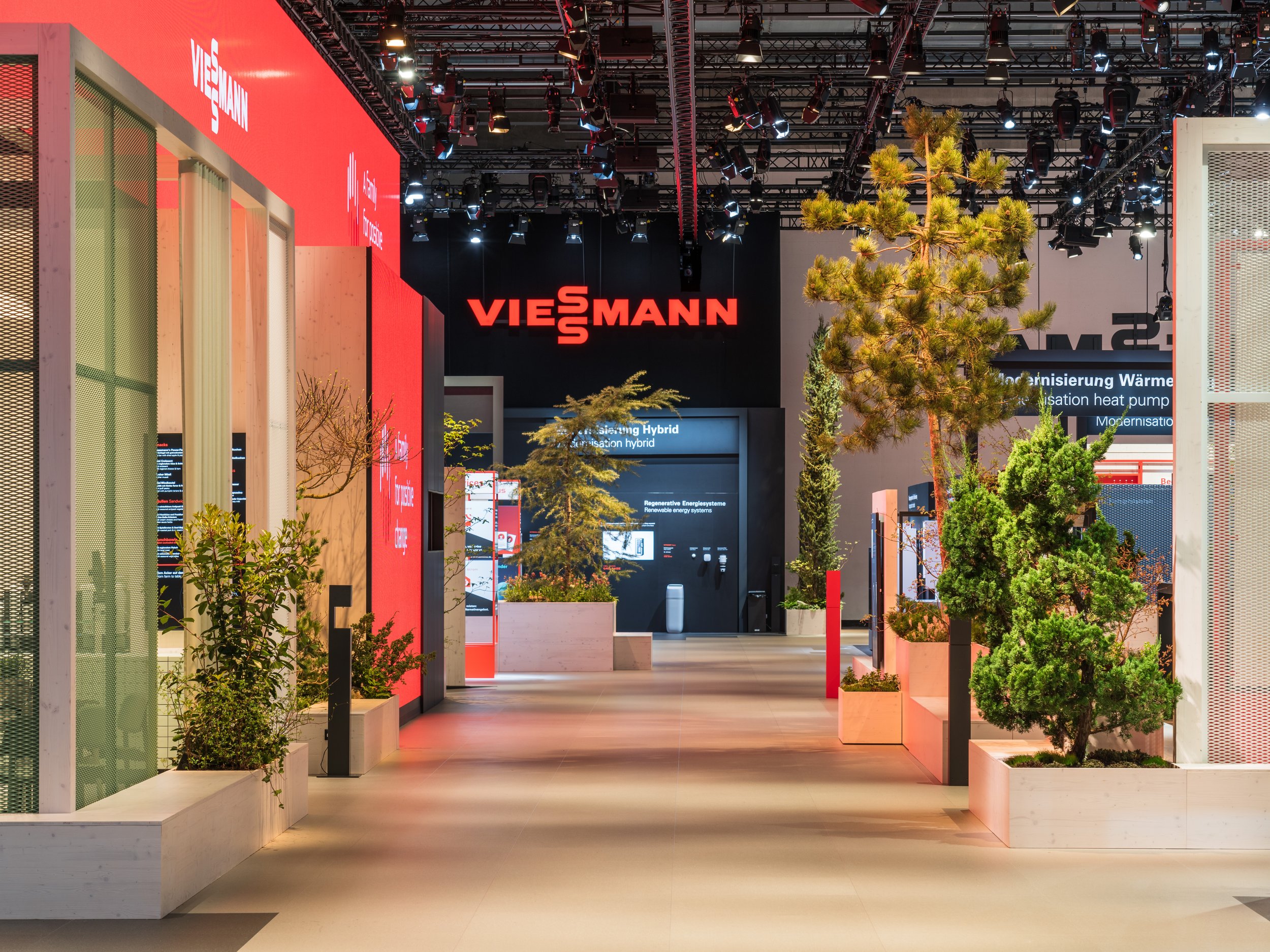 Indoor exhibition space with plants in white planters, illuminated signs, and a large red and black sign reading "VIESMANN" in the background.