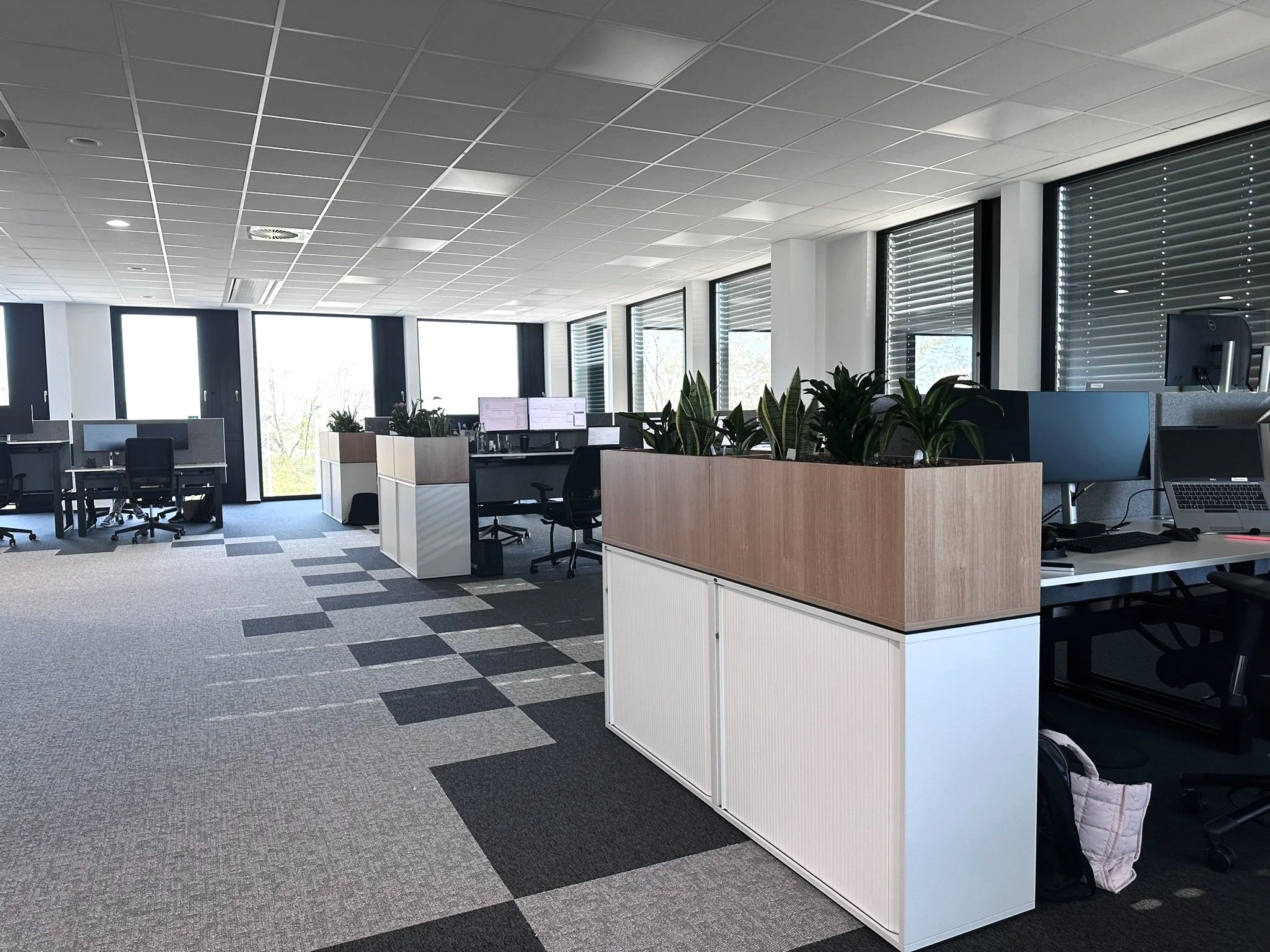 An empty modern office space with work desks, chairs, and computer monitors, illuminated by large windows and skylights.