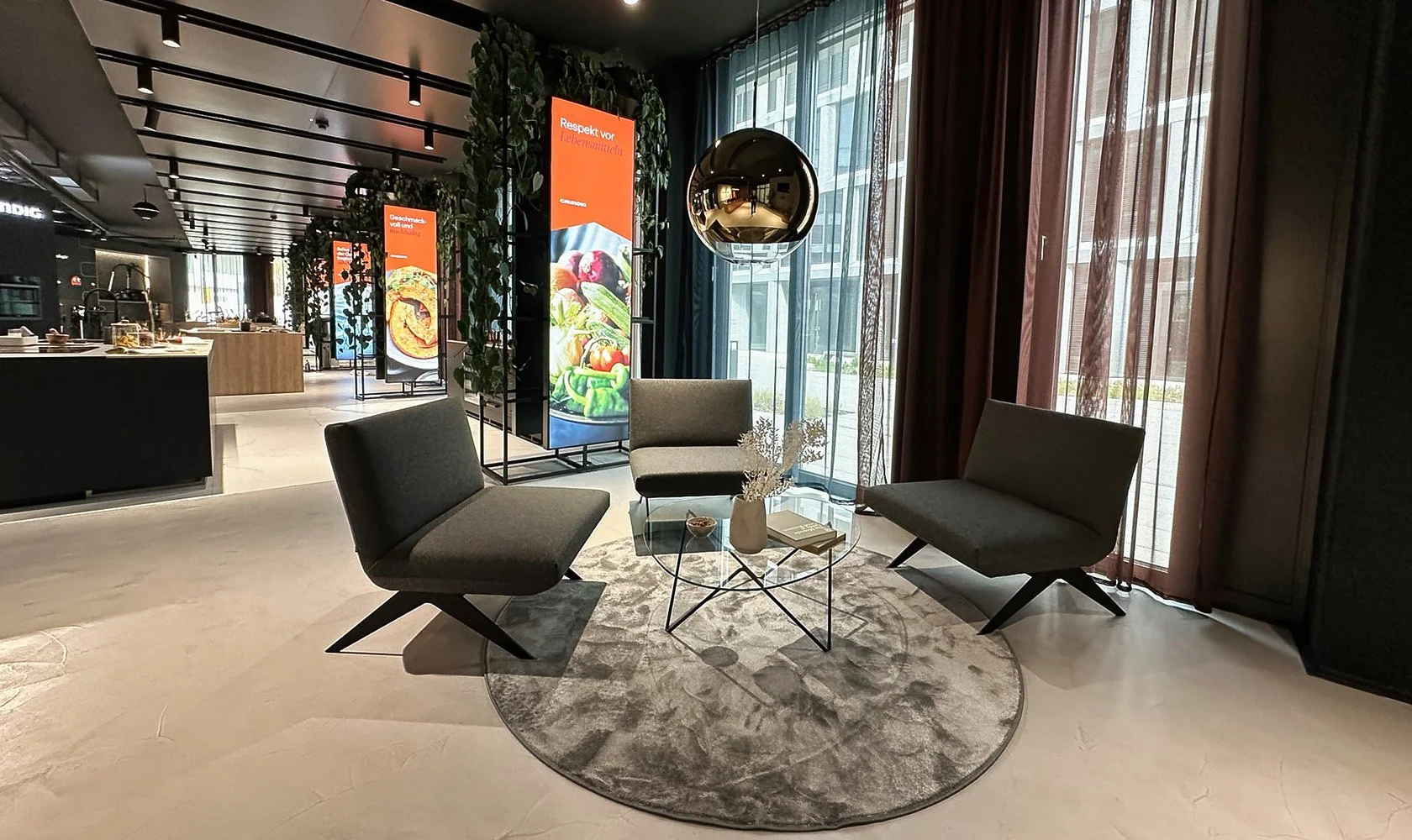 Modern lobby area with gray chairs around a glass coffee table, decorative vases, large windows with curtains, and illuminated display boards showing food images.