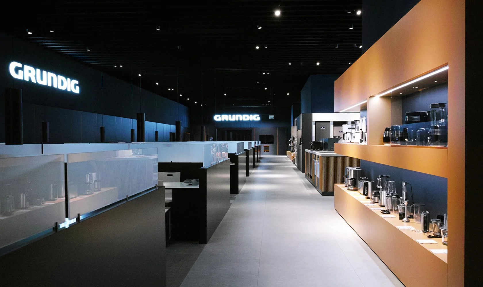 Empty store display area with coffee makers and kitchen appliances on orange shelves, illuminated signs that say "GRUNDIG" in the background.