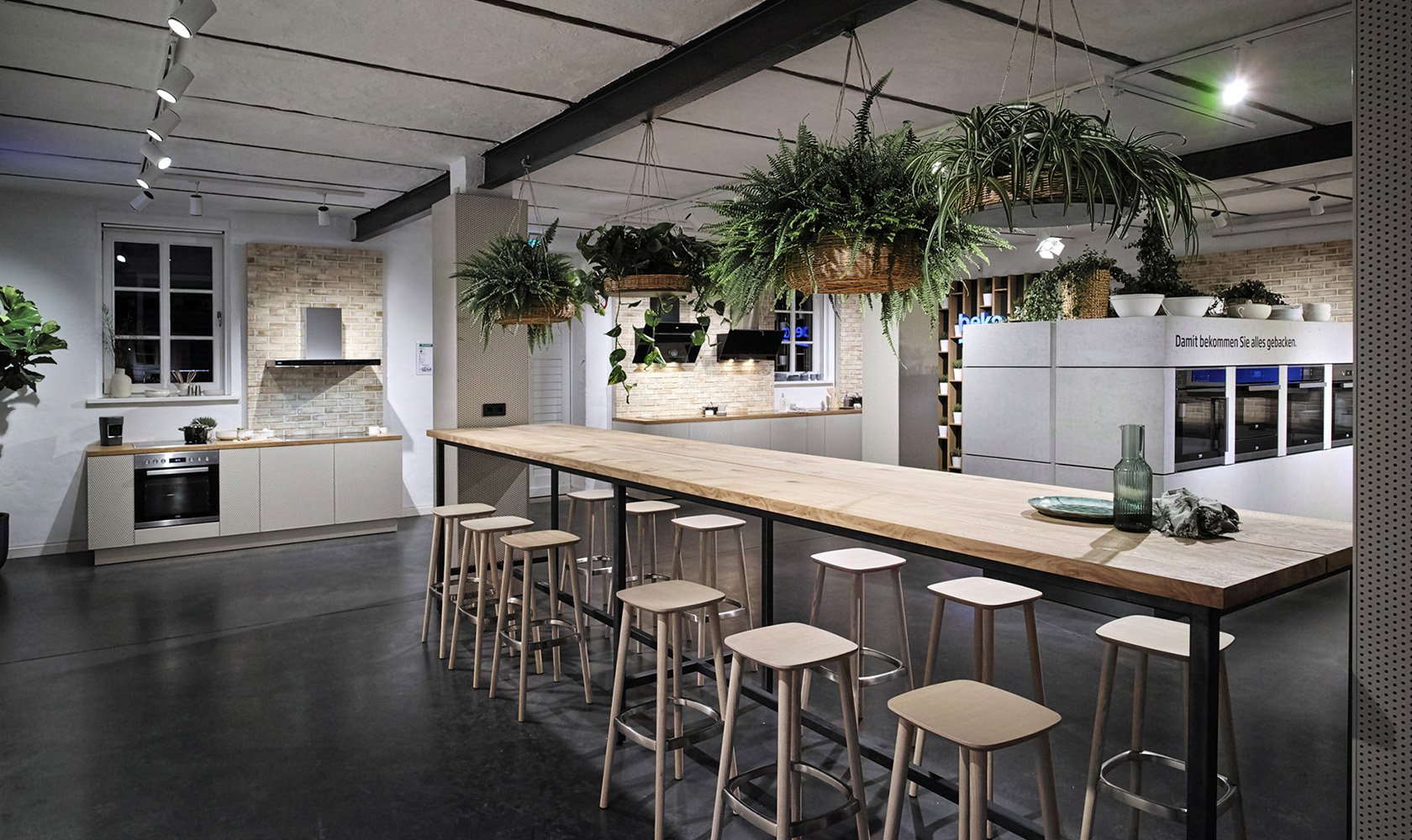 Modern kitchen with a long wooden table, hanging plants, and white cabinetry, illuminated by track lighting and featuring a brick wall behind the stove.