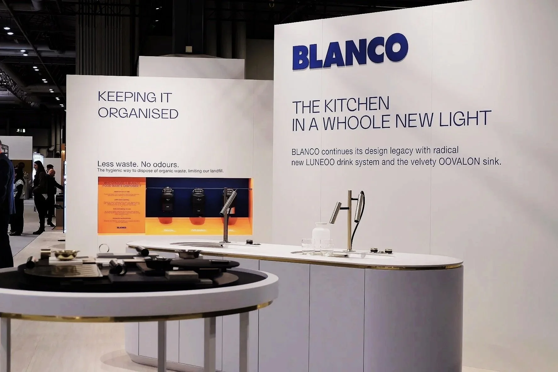 Exhibition booth displaying kitchen fixtures, with a large white wall featuring the brand name 'BLANCO' and promotional text about kitchen design in a new light. A countertop with a sink, faucet, and bottles, and an area highlighting waste disposal solutions.