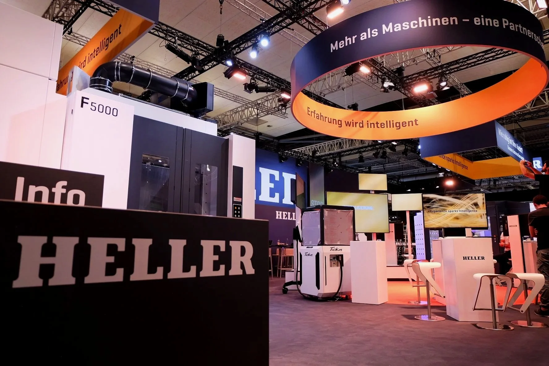 Trade show booth for Heller with various machines, screens displaying advertising, and hanging banners in German promoting smarter machinery and intelligent experiences