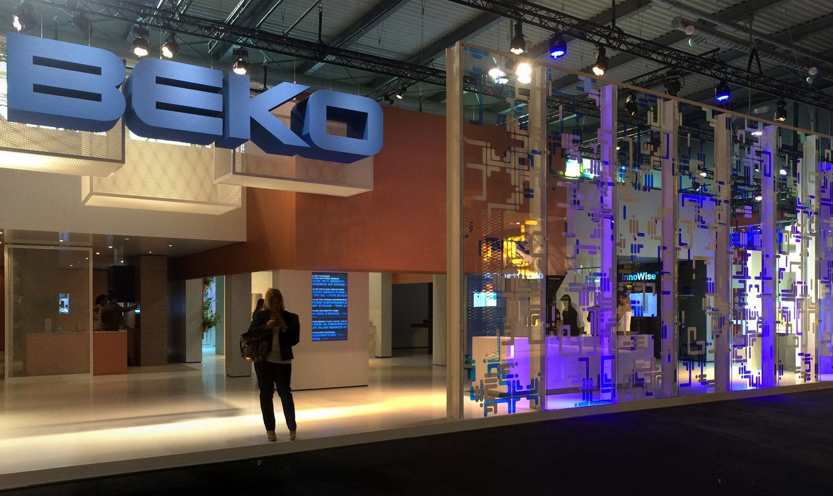 Trade show booth with a large blue 'BEKO' sign, modern design elements, and illuminated displays, with people inside and outside the booth.