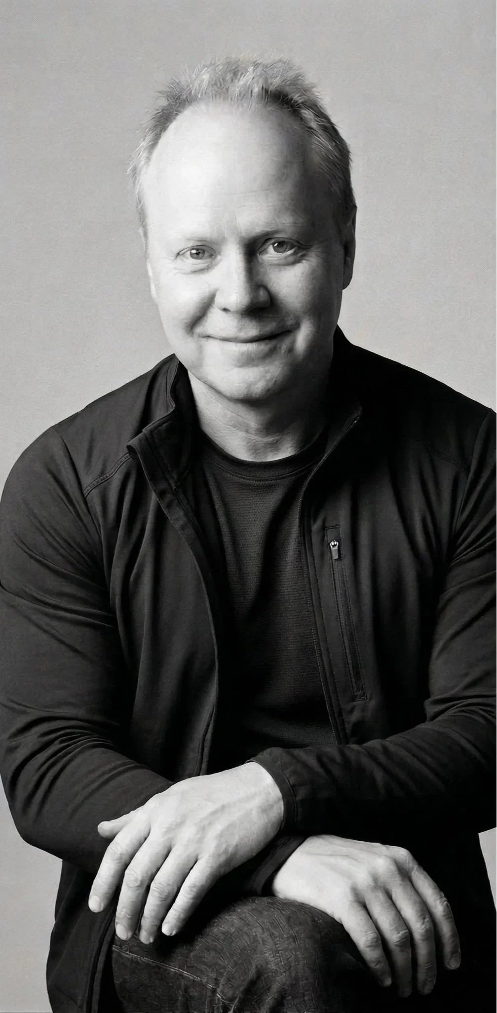 Black and white portrait of a smiling middle-aged man with short hair, wearing a black jacket and sitting with his hands resting on a chair.