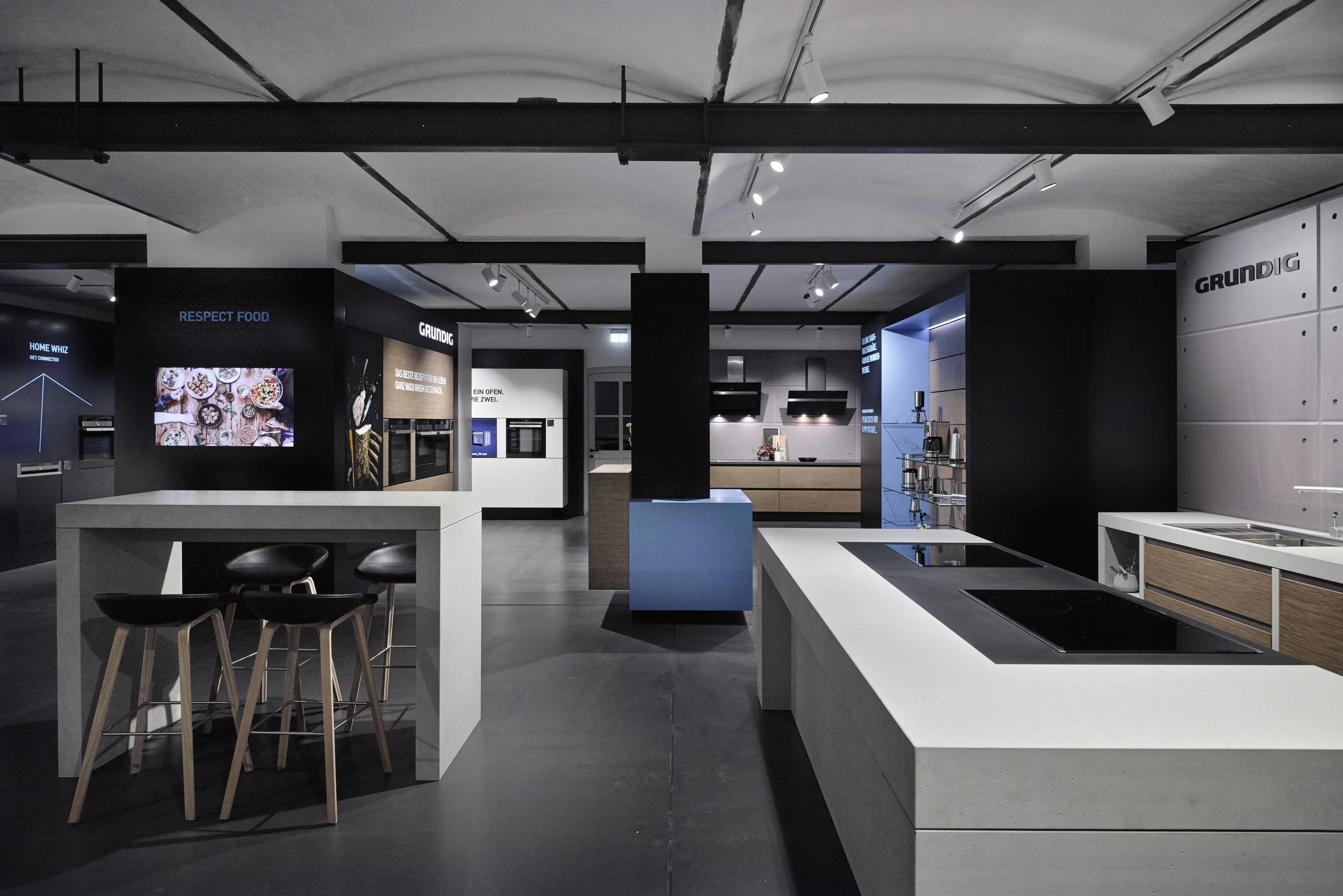 A modern kitchen showroom with black, white, and wooden accents. Features include a white island with induction cooktops, a high white table with three black stools, and various kitchen appliances on display. The background shows shelves with kitchen products and illuminated display panels.