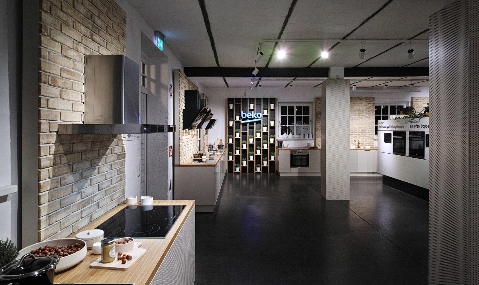 Modern kitchen display with brick walls, wooden furniture, and a shelving unit with a illuminated 'beko' sign and potted plants.