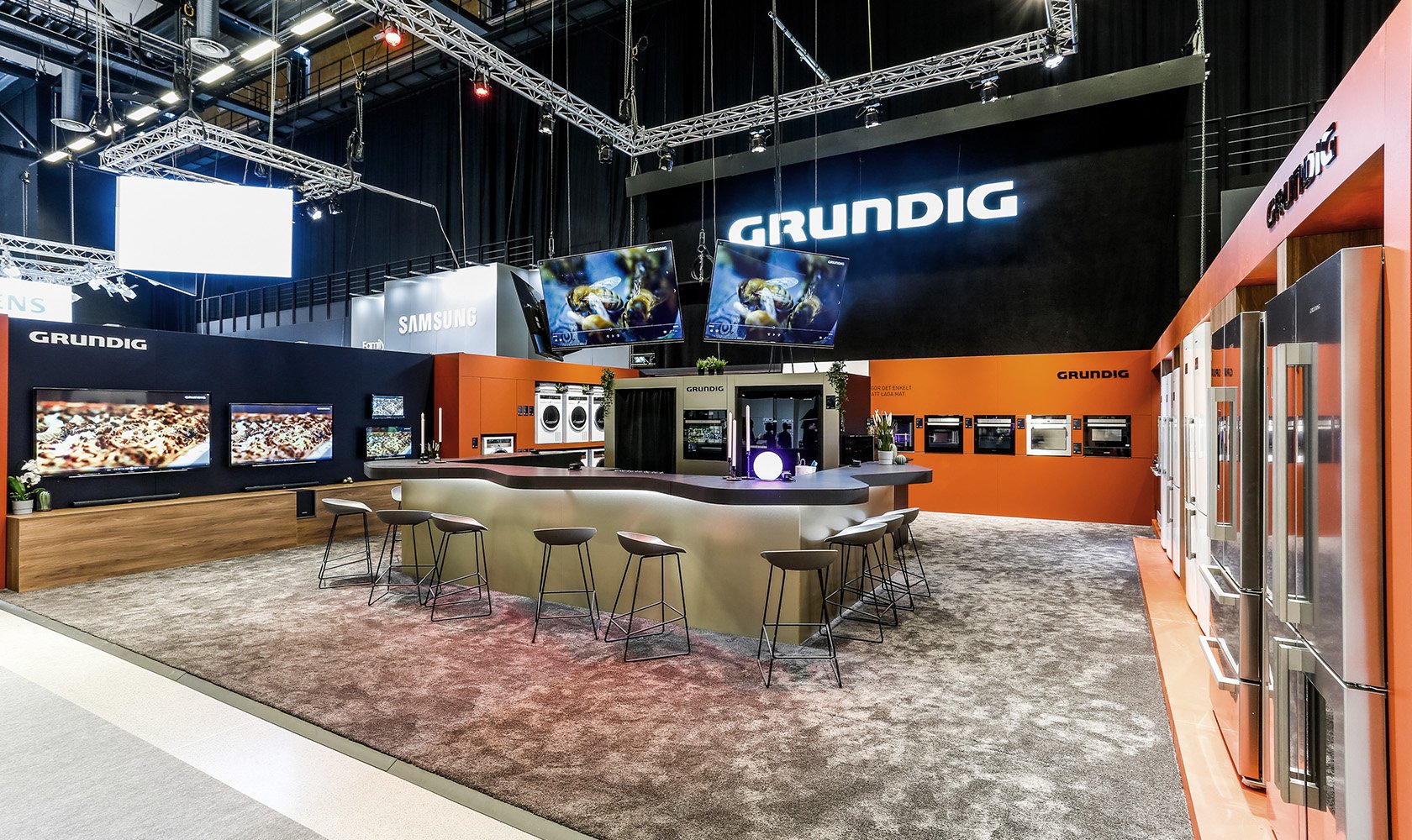 Trade show booth for Grundig with appliances, multiple screens, and seating area, in an indoor exhibition space.