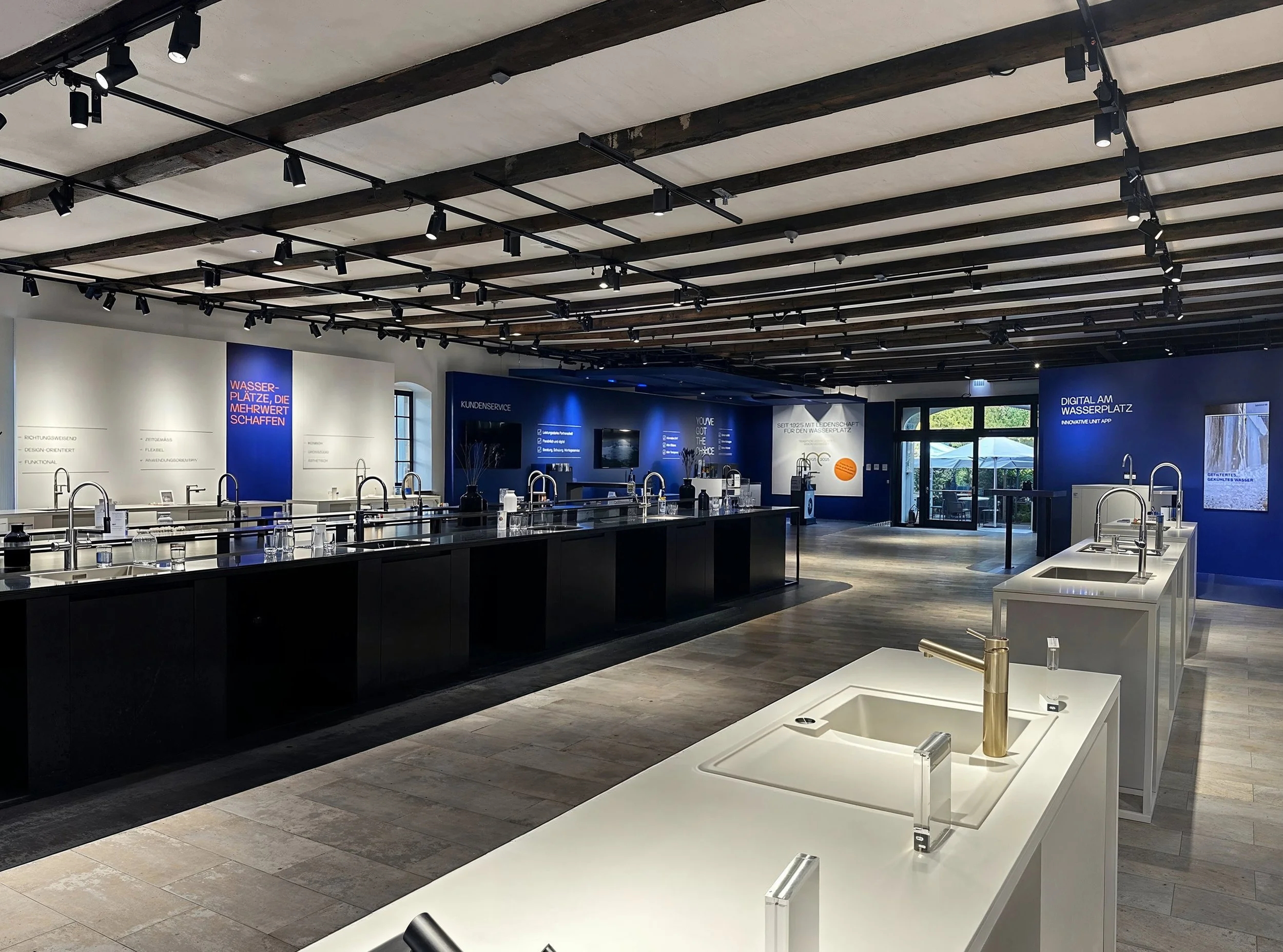 Showroom display of kitchen sinks and faucets with illuminated blue and white walls, signs in German, and large windows letting in natural light.