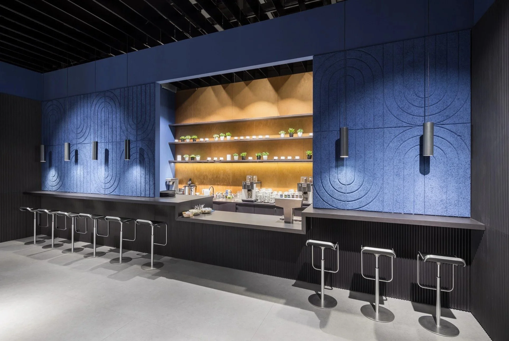 Modern coffee bar with black counter, five bar stools, blue textured walls, and shelves with potted plants and cups.