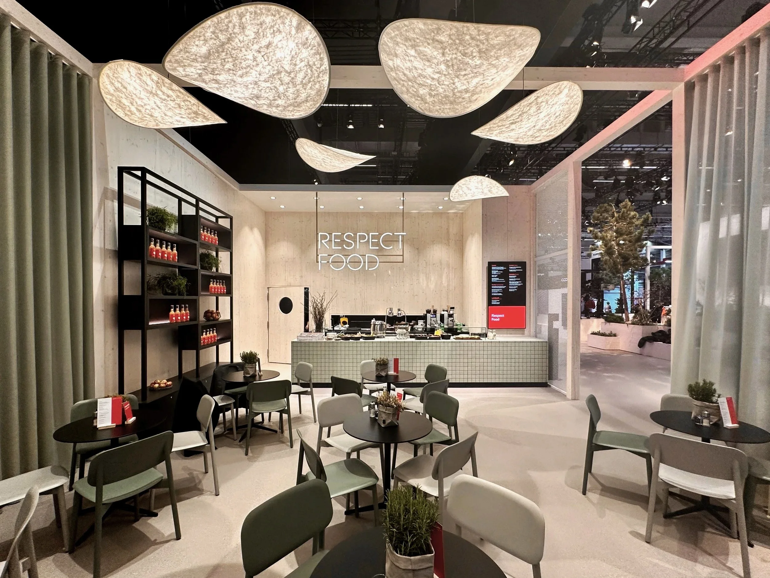 Modern dining area with tables and chairs, a display shelf with bottles, a counter with food and drink items, and a sign that reads 'Respect Food' in a well-lit, contemporary setting.