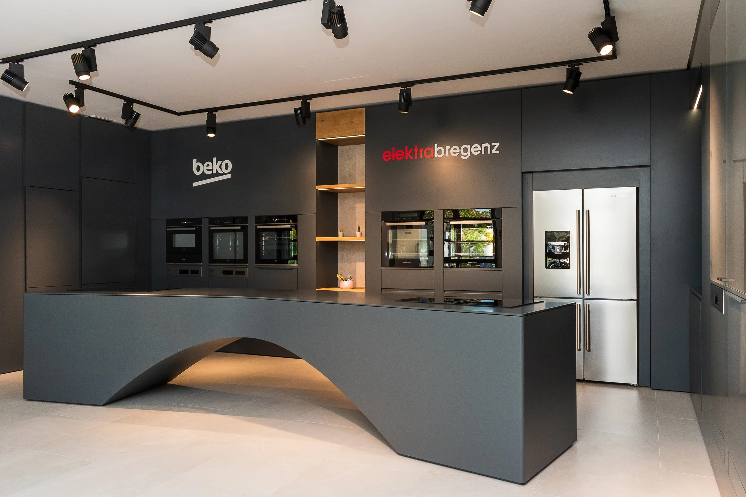 Modern kitchen showroom with black cabinets, built-in ovens, and a large metallic refrigerator. A unique gray island counter with a curved design is in the foreground. Wall signs display the brands 'beko' and 'elektrabregenz,