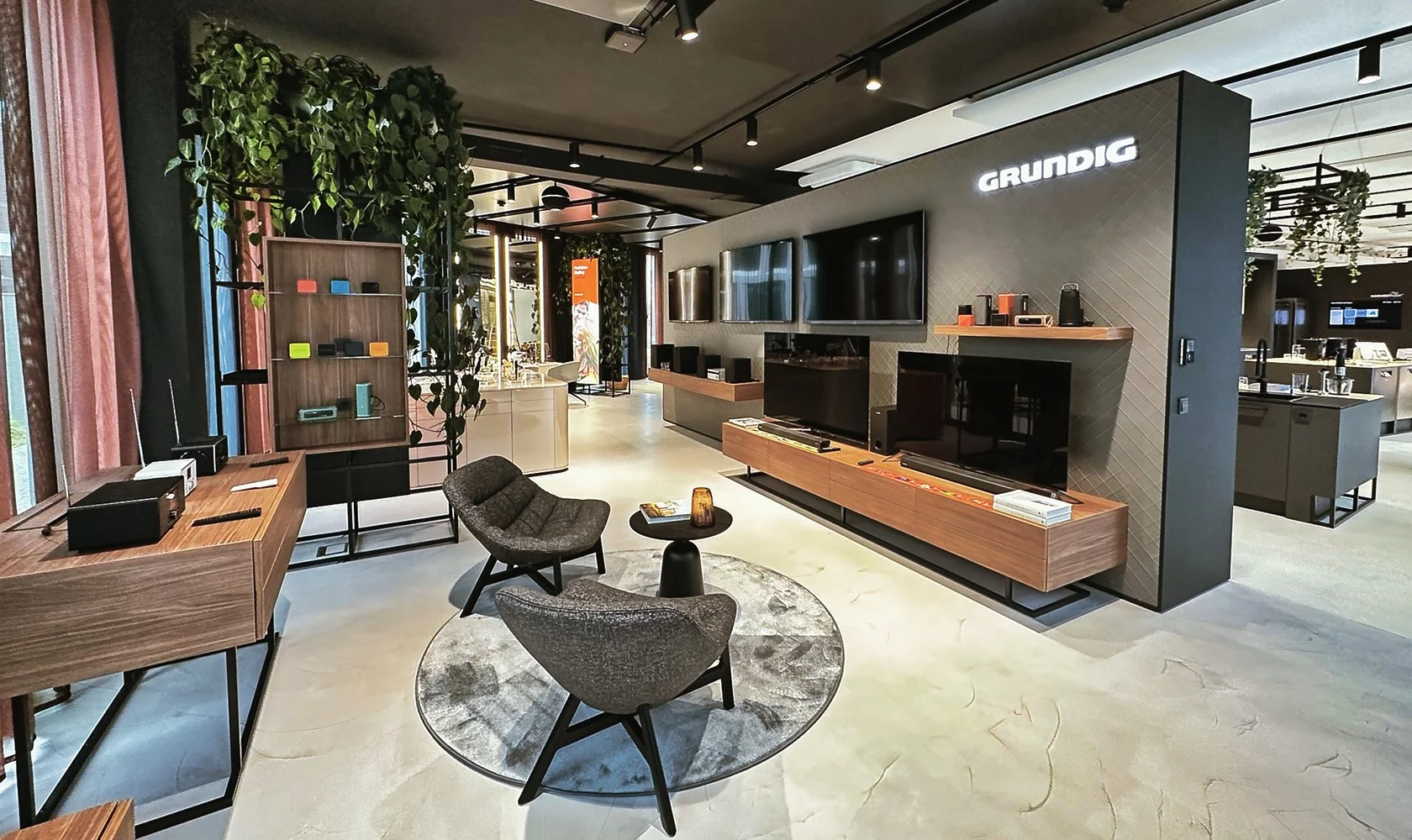 Modern electronics showroom with TVs, speakers, and home entertainment displays, featuring a seating area with two chairs and a small table, and product shelves on the walls.