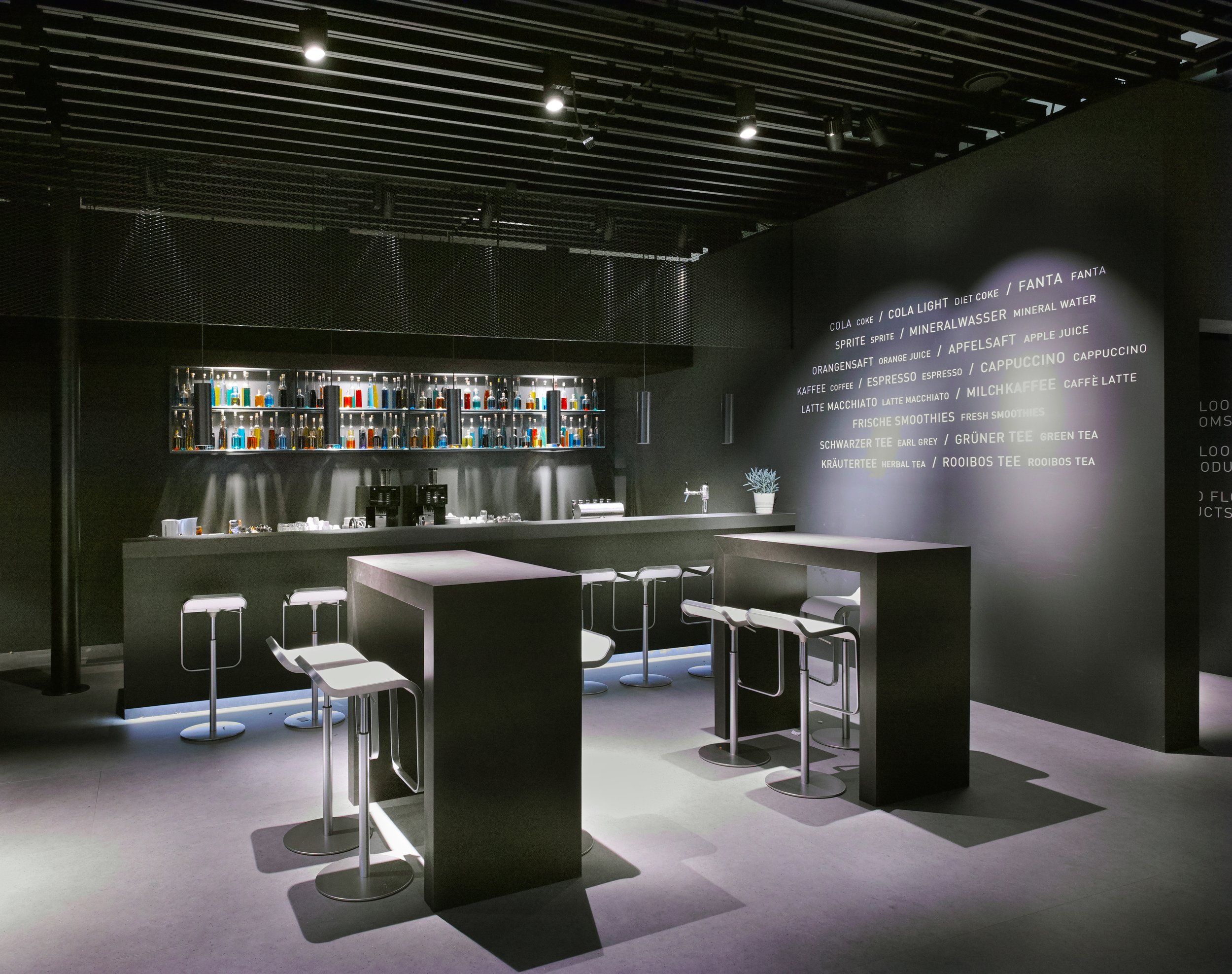 A modern, minimalist café or bar with black walls and furniture. There are high tables with stools, a black counter with coffee machines, and shelves displaying colorful bottles. On the wall, there is a menu listing drinks such as cola, orange juice, coffee, teas, smoothies, and water.