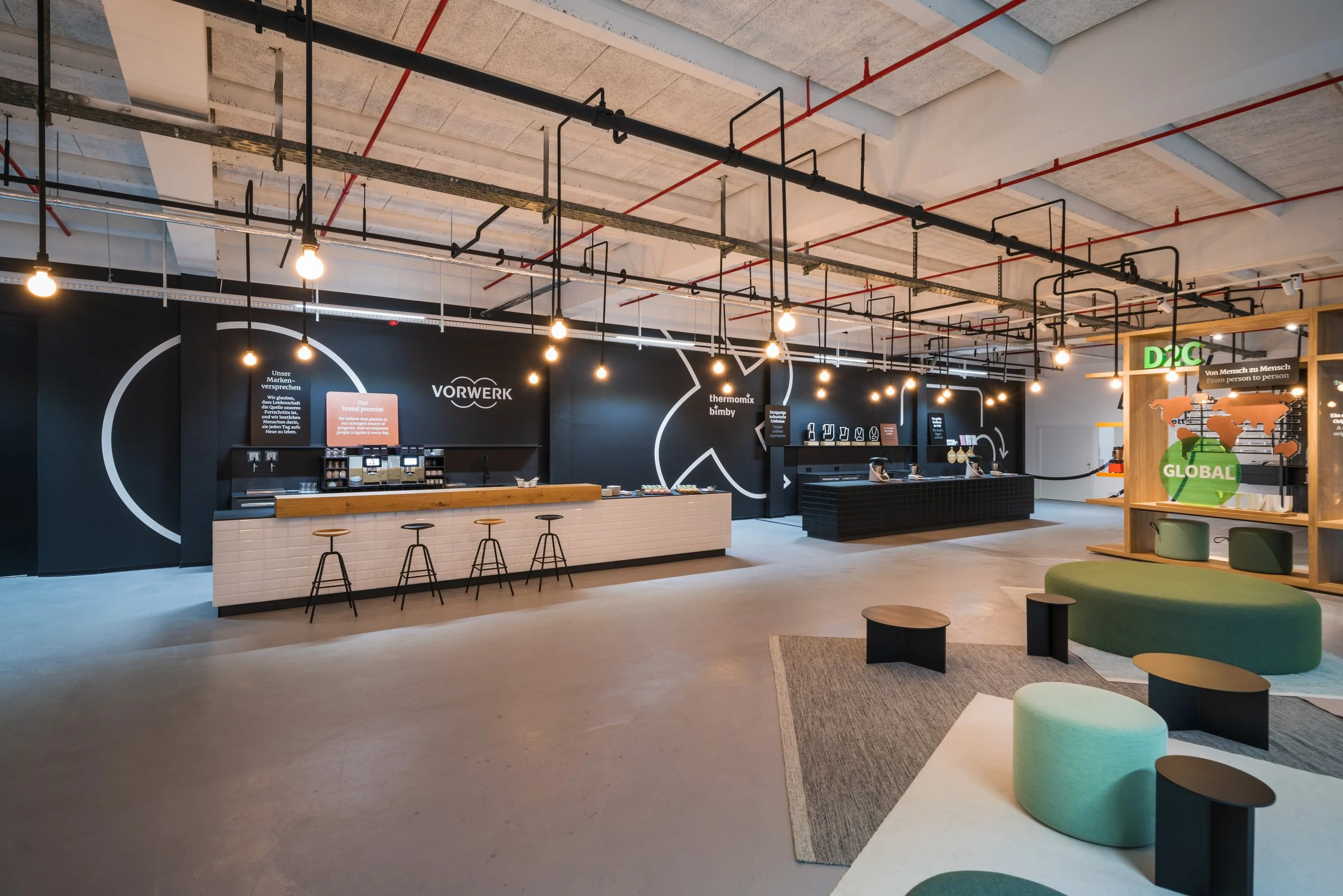 Modern coffee shop with black walls, illuminated by hanging bulbs, featuring a long white counter with stools, seating area with green and black furniture, and a display shelf with global-themed decor.