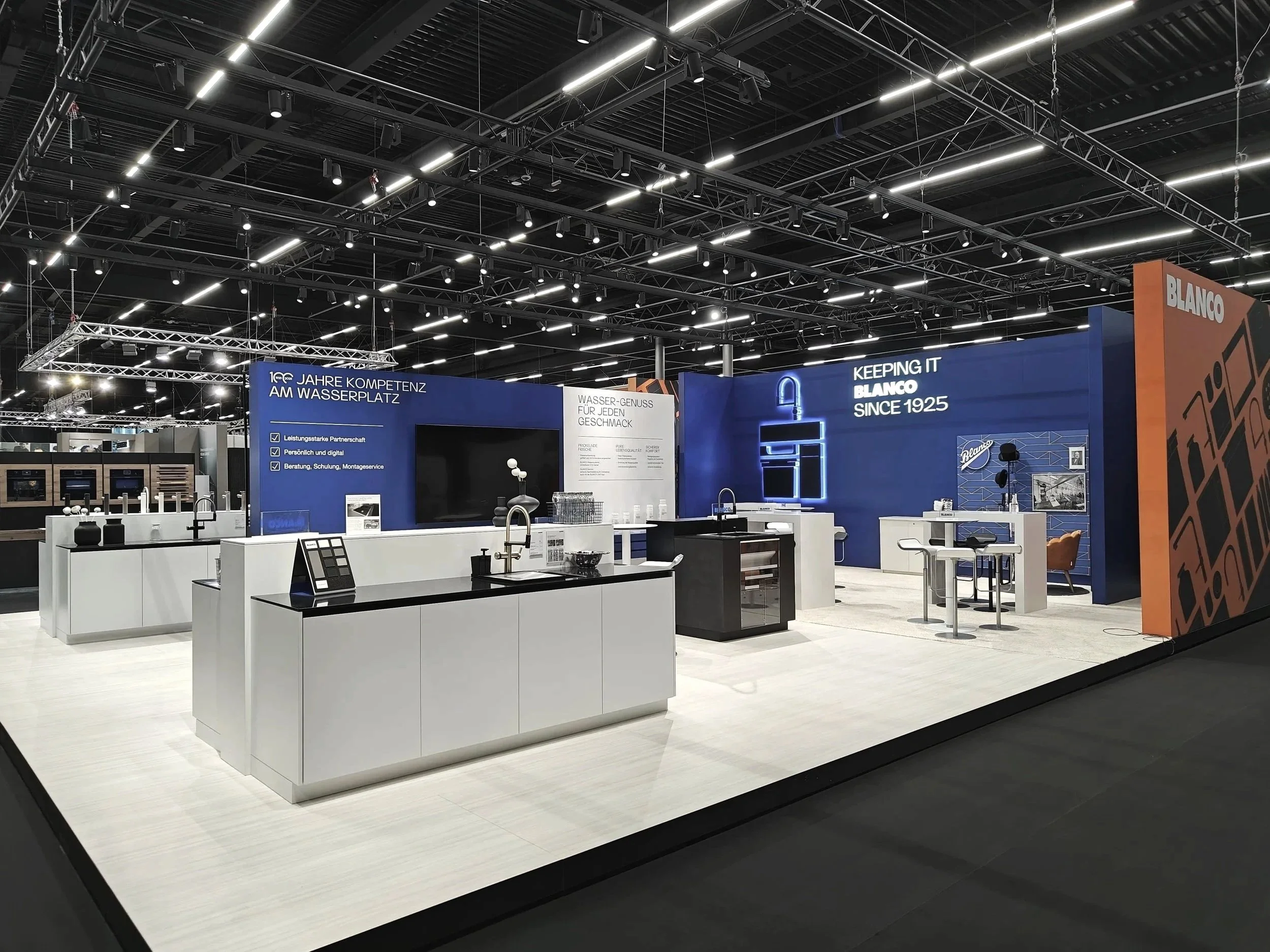 A trade show booth with modern kitchen fixtures and appliances, featuring blue and white walls, a large TV, and neon signage reading 'KEEPING IT BLANCO SINCE 1925'.