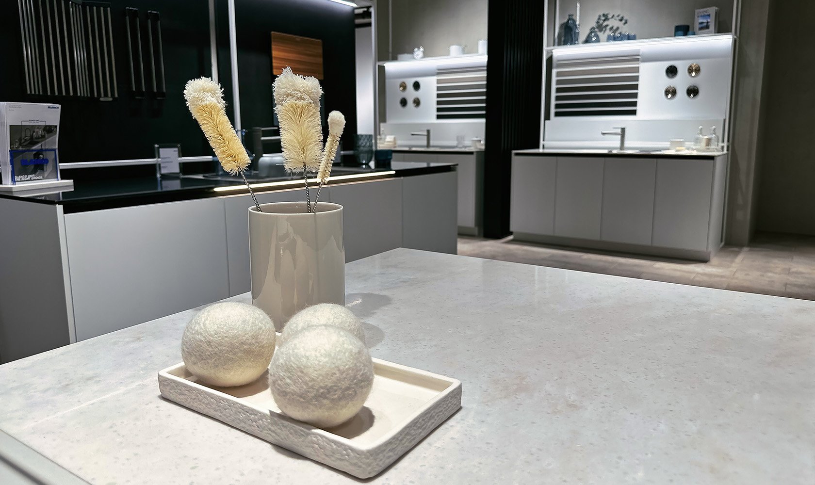 A modern kitchen with a white countertop. On the counter, there is a white tray holding three round, textured white balls. Behind the tray, there is a beige vase with dried pampas grass. The background features white cabinets, black walls, and kitchen equipment.