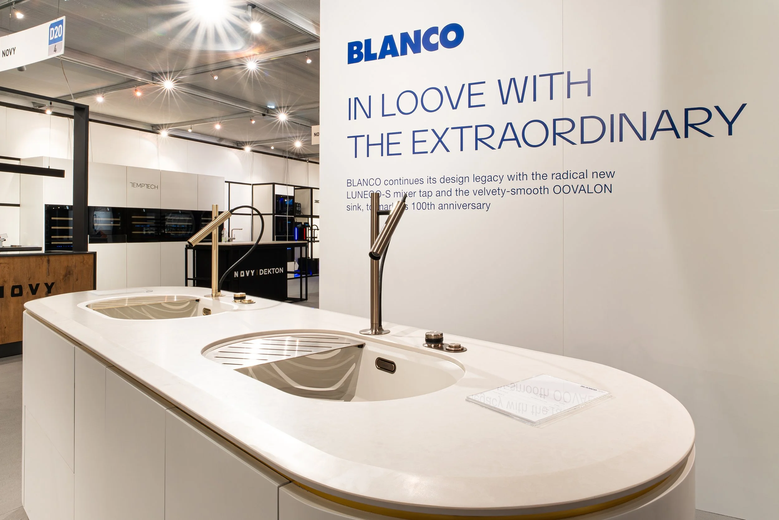 Display of a modern kitchen sink with a curved white countertop, two integrated stainless steel sinks, and dual faucets, at a trade show or exhibition.