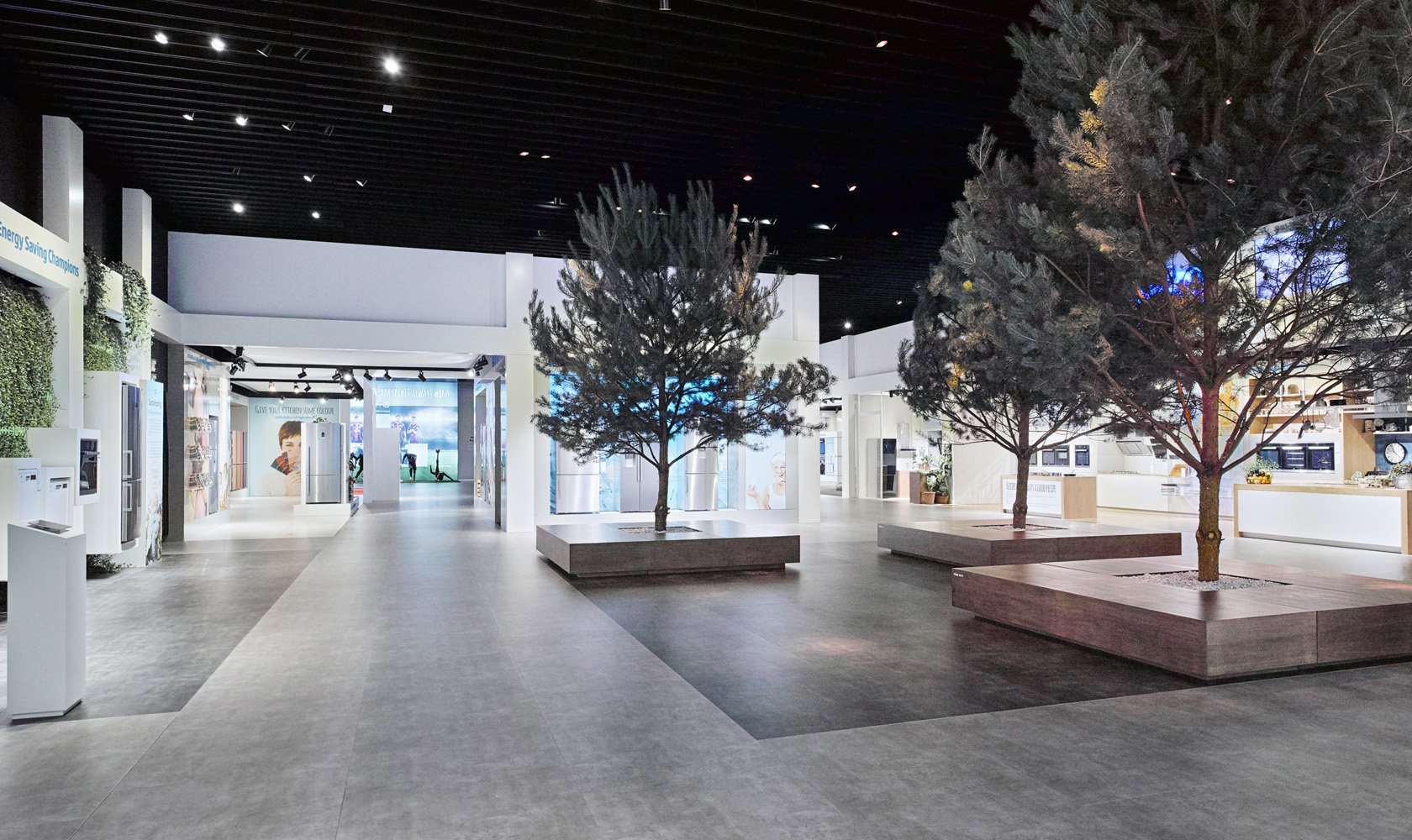 Interior of a modern shopping mall or store with trees in planters, display areas, and a food court or seating area in the background.