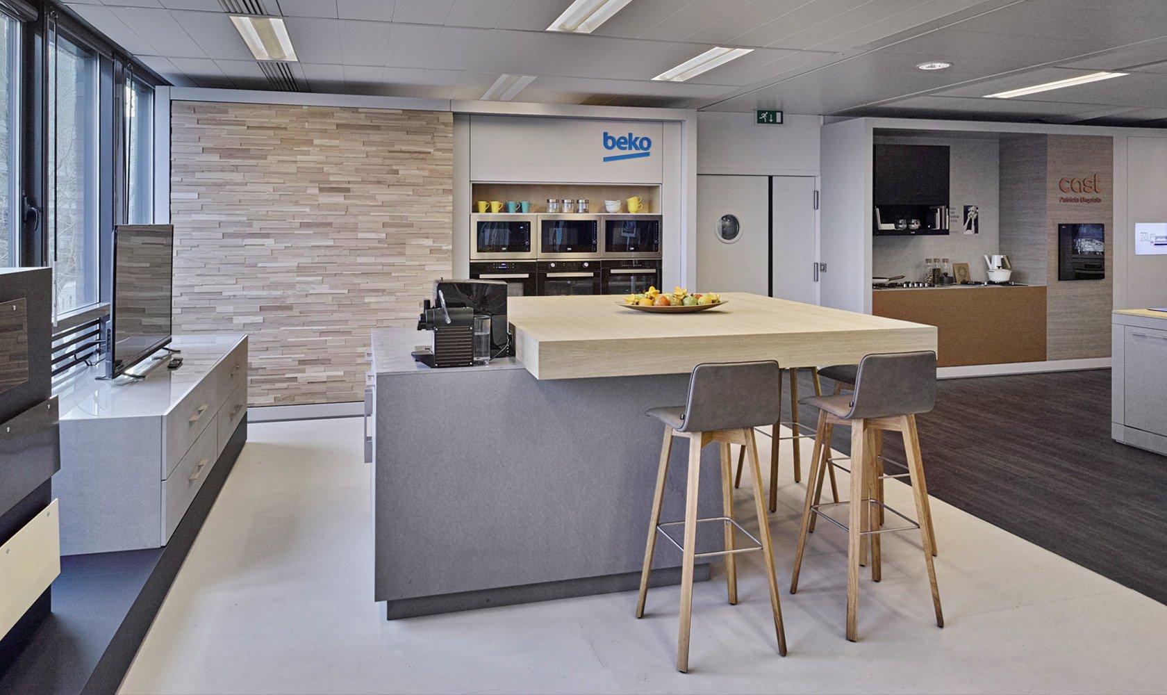 Modern office kitchenette with a kitchen island, seating area, and appliances including microwaves and coffee maker, with a large window on the left side.