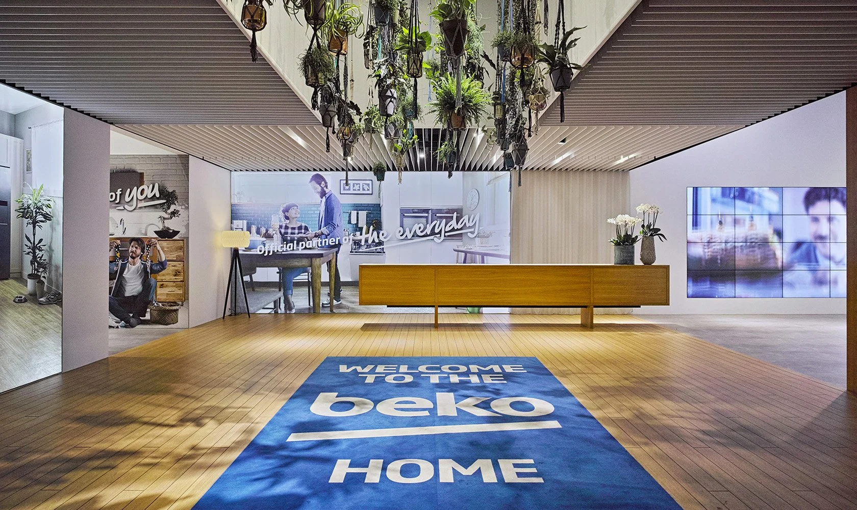 Reception area with wooden flooring and a large blue rug that says "Welcome to the beko home." The space has hanging plants on the ceiling, a wooden bench, a wall with images and text, and a digital screen displaying a scene.