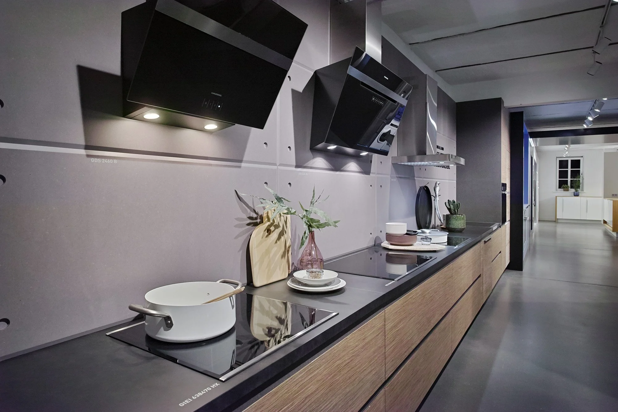 Modern kitchen with black cooktop, wooden cabinets, gray backsplash, stainless steel range hood, and decorative items like bowls, plants, and cutting boards.