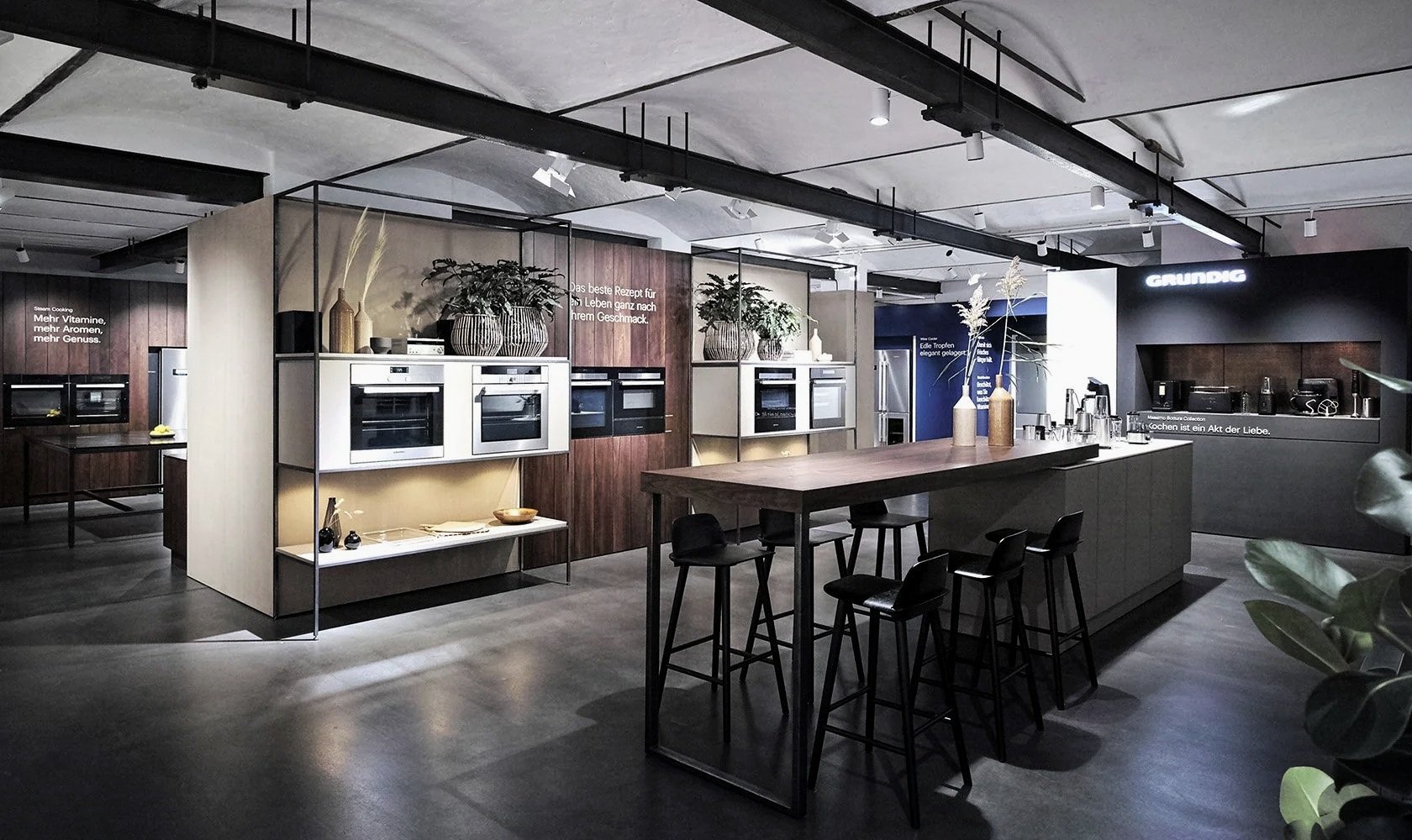 Modern kitchen showroom with built-in ovens, a large central island with bar stools, and decorative plants and vases, featuring a sleek, contemporary design.