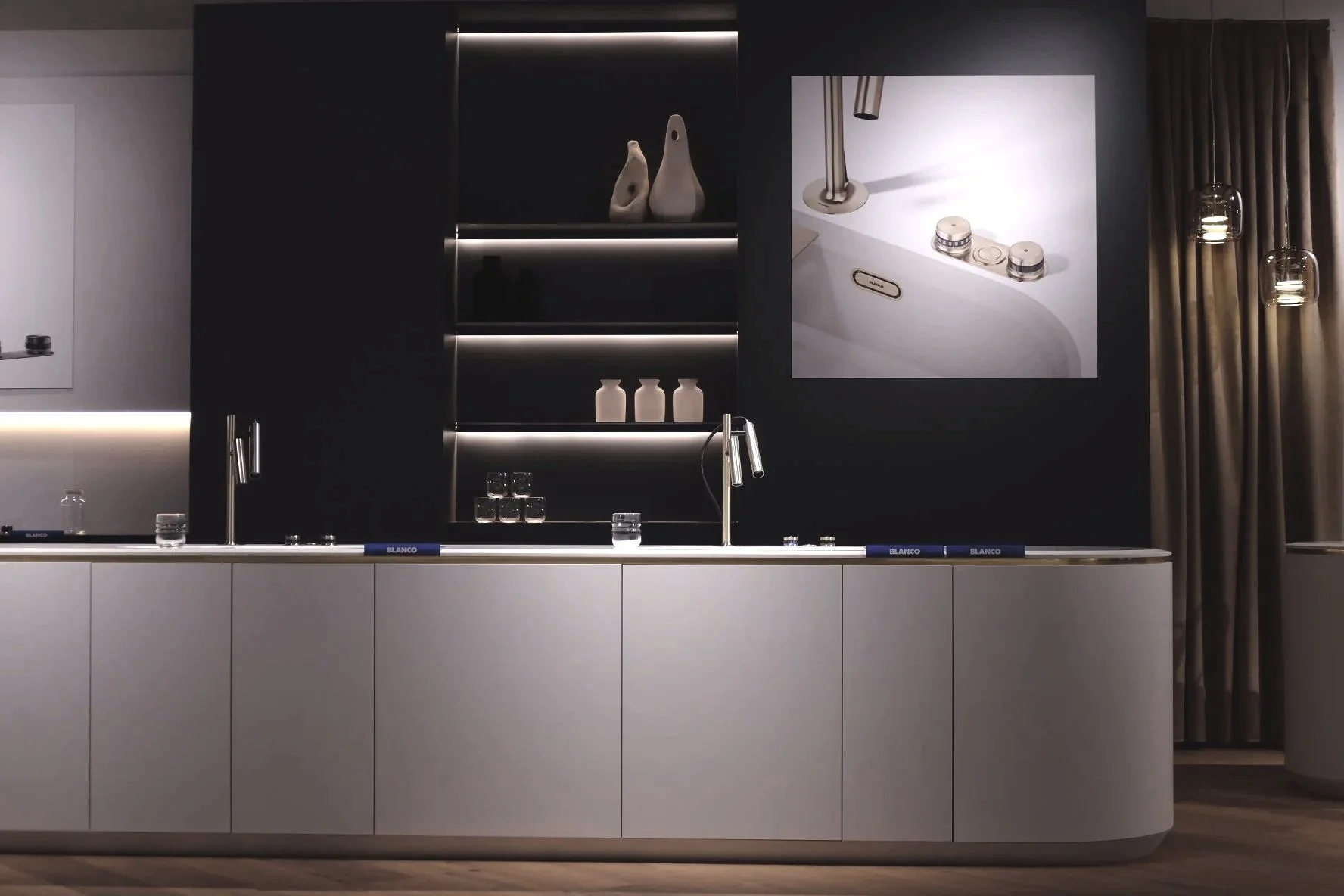 Modern minimalist kitchen with white lower cabinets, black upper cabinets, and a black backsplash with built-in lighting. The countertop has small glasses and books, a sleek silver faucet, and a large poster on the wall showing a close-up of a sink with a faucet and knobs. Two pendant lights hang to the right, and beige curtains cover a window on the far right.