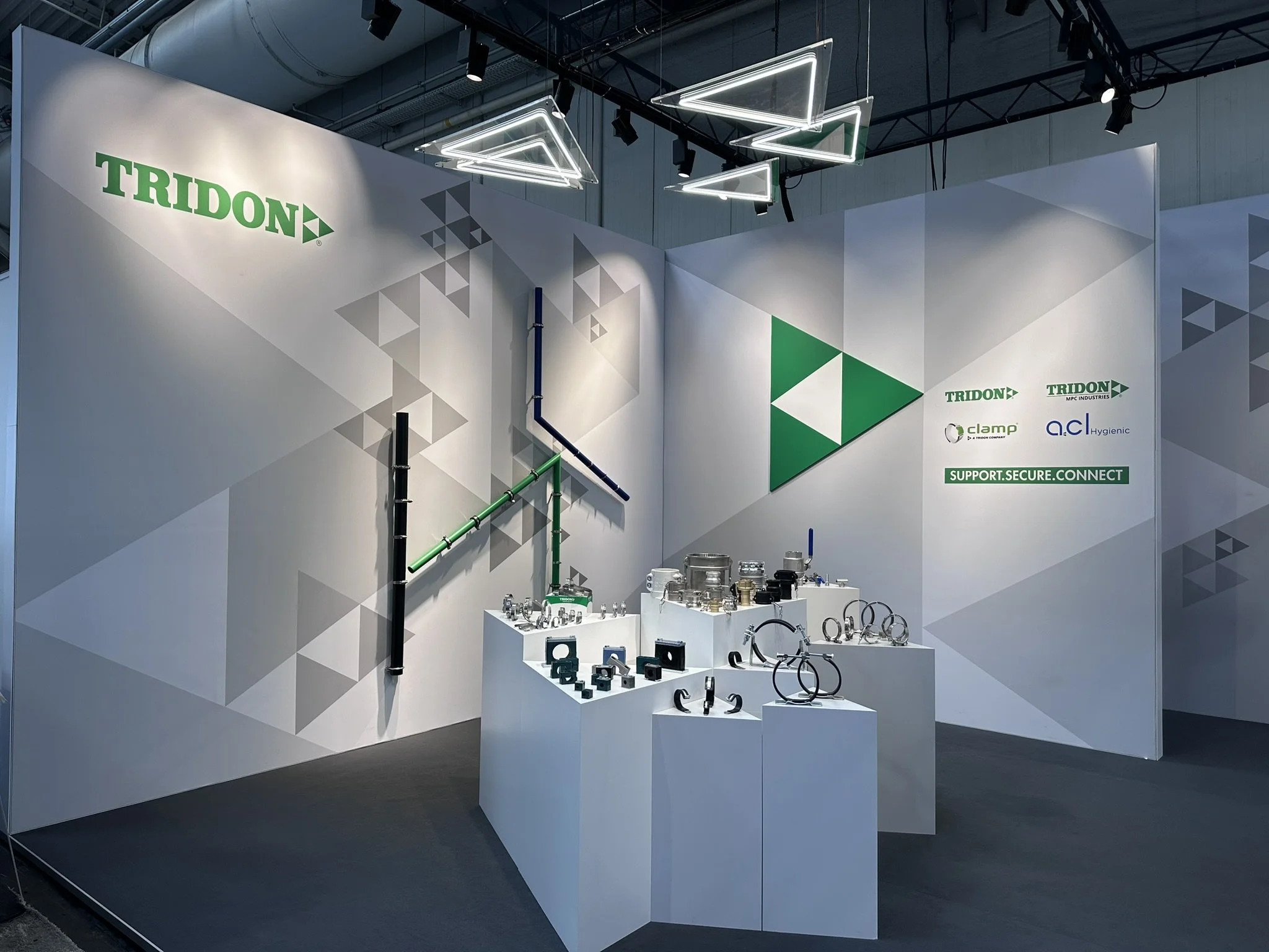 Trade show booth with the logo 'TRIDON' displayed on the wall, showcasing various industrial pipes, fittings, and equipment on white display tables, with lighting fixtures hanging from the ceiling.
