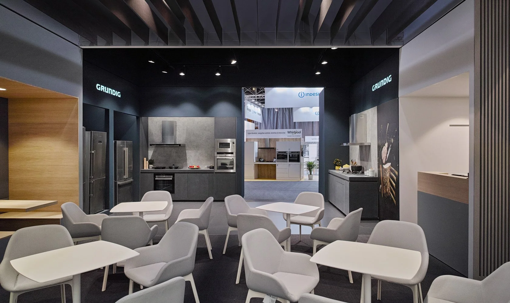 Modern kitchen display area with grey cabinetry, stainless steel appliances, and a black backdrop. In the foreground, there are white chairs and tables for seating.