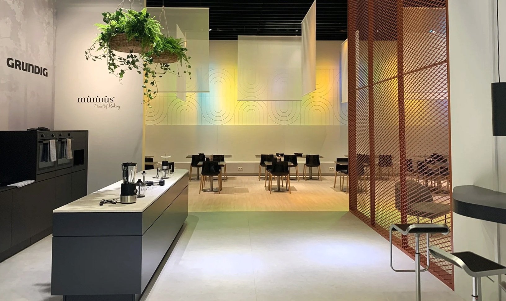 Modern cafe with black water dispensers, a white marble counter with kitchen appliances, black chairs around tables, a decorative wall with lighting, and an orange metal partition